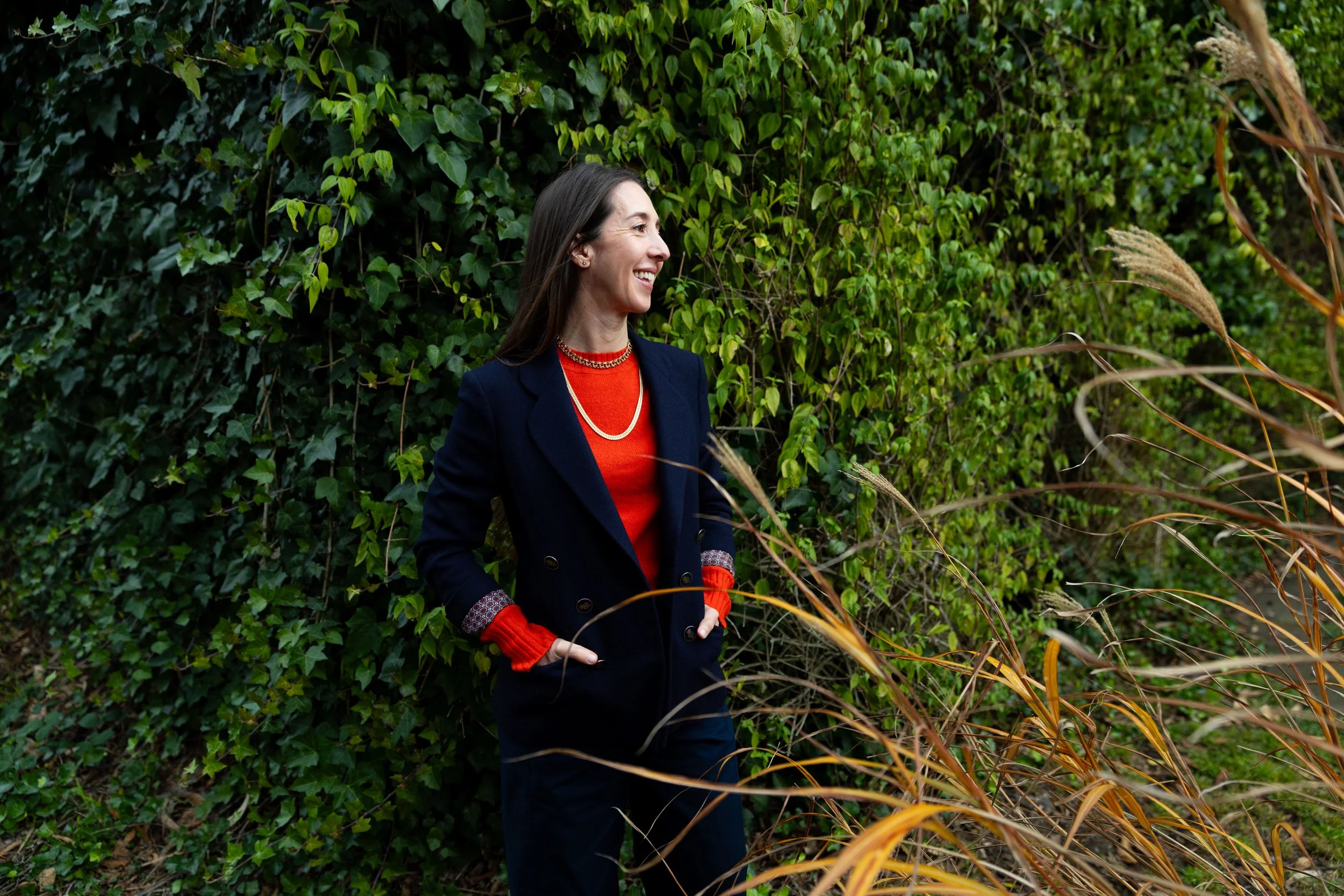 A woman standing outdoors, smiling and looking to the side, surrounded by green foliage and brown grasses.