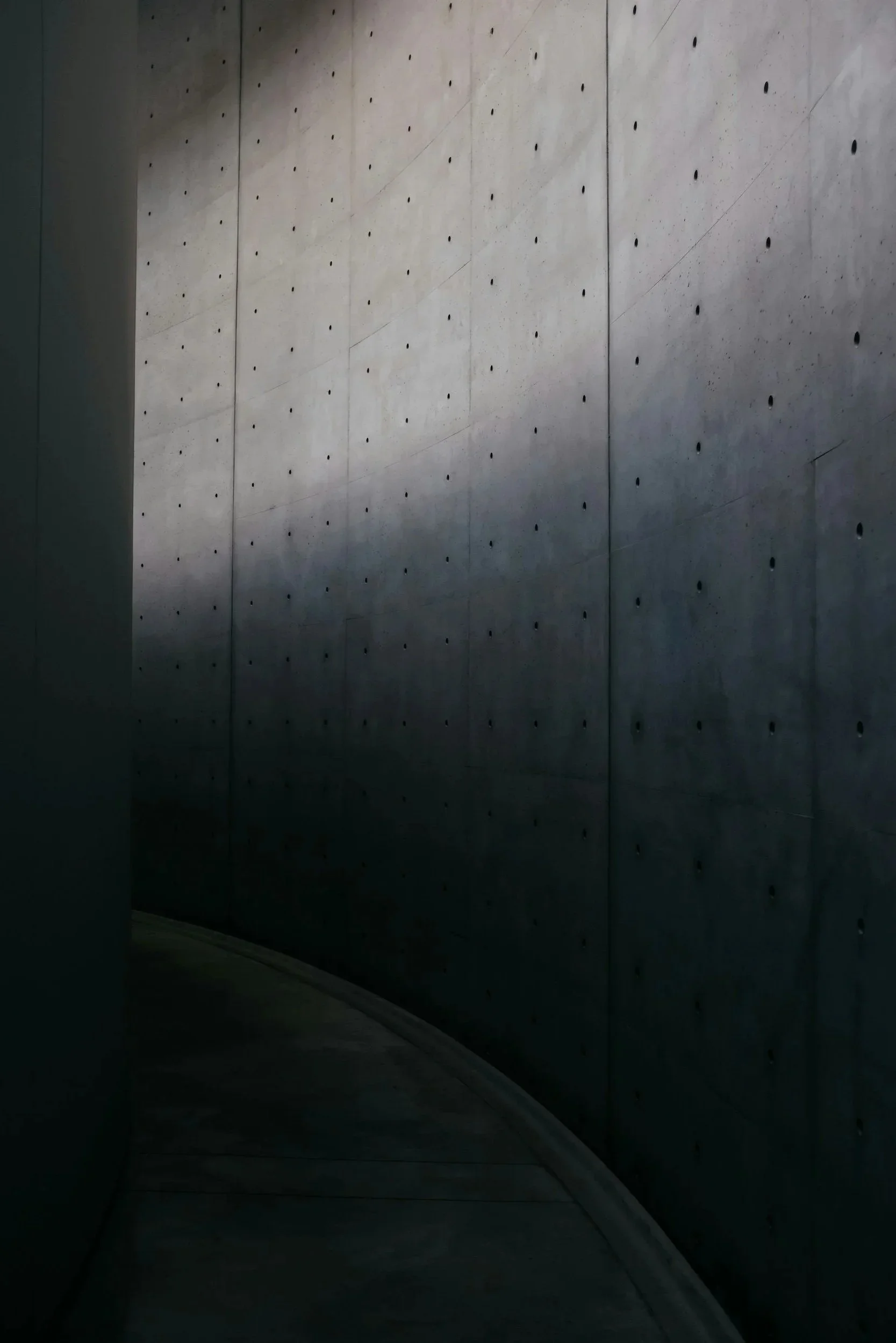 A curved concrete wall with small evenly spaced holes and a narrow shadow cast on it.