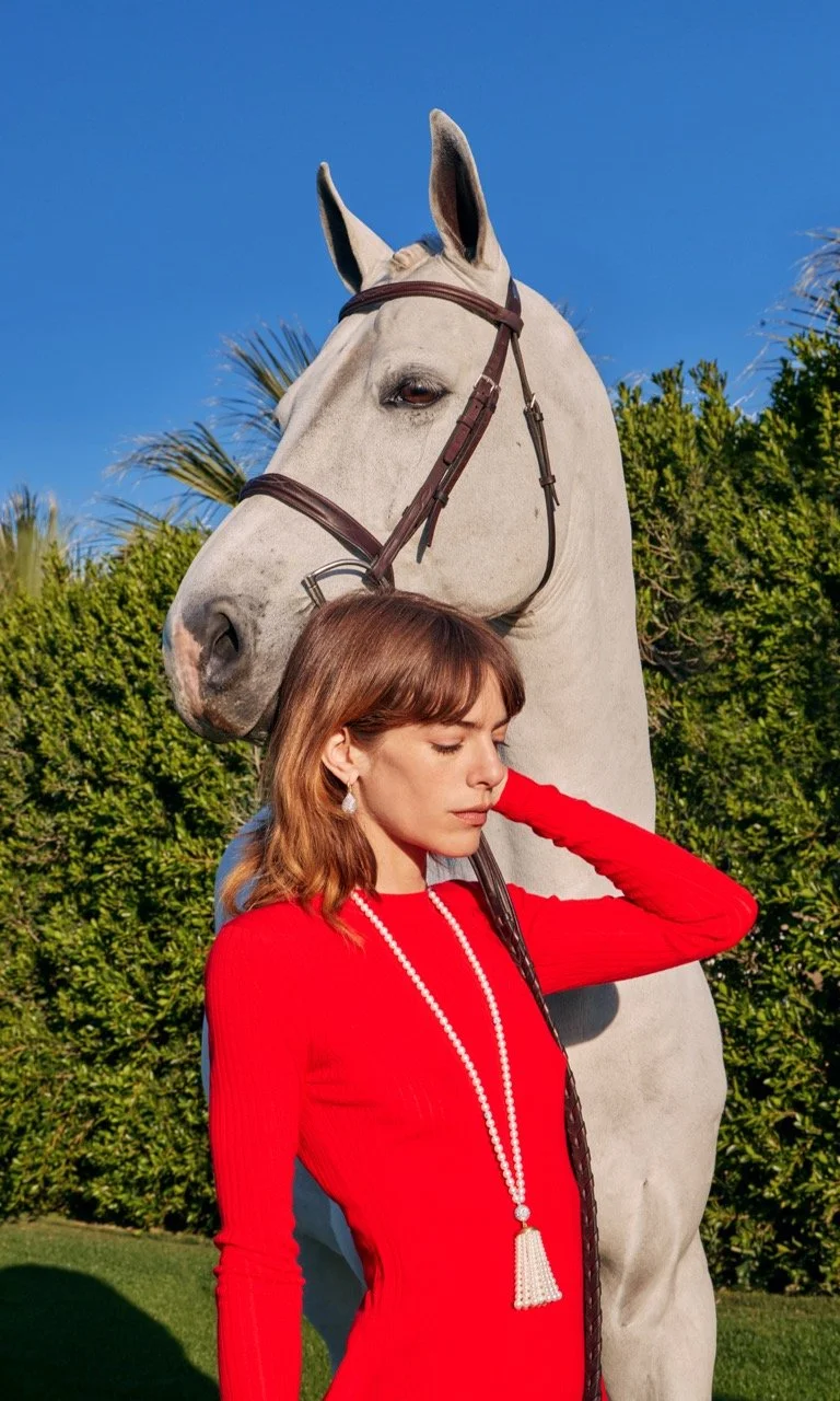 A woman in a red dress with a pearl necklace standing outdoors with a white horse behind her. The woman has brown hair and is rubbing her neck with her left hand, with her eyes closed. The background includes greenery and a clear blue sky.