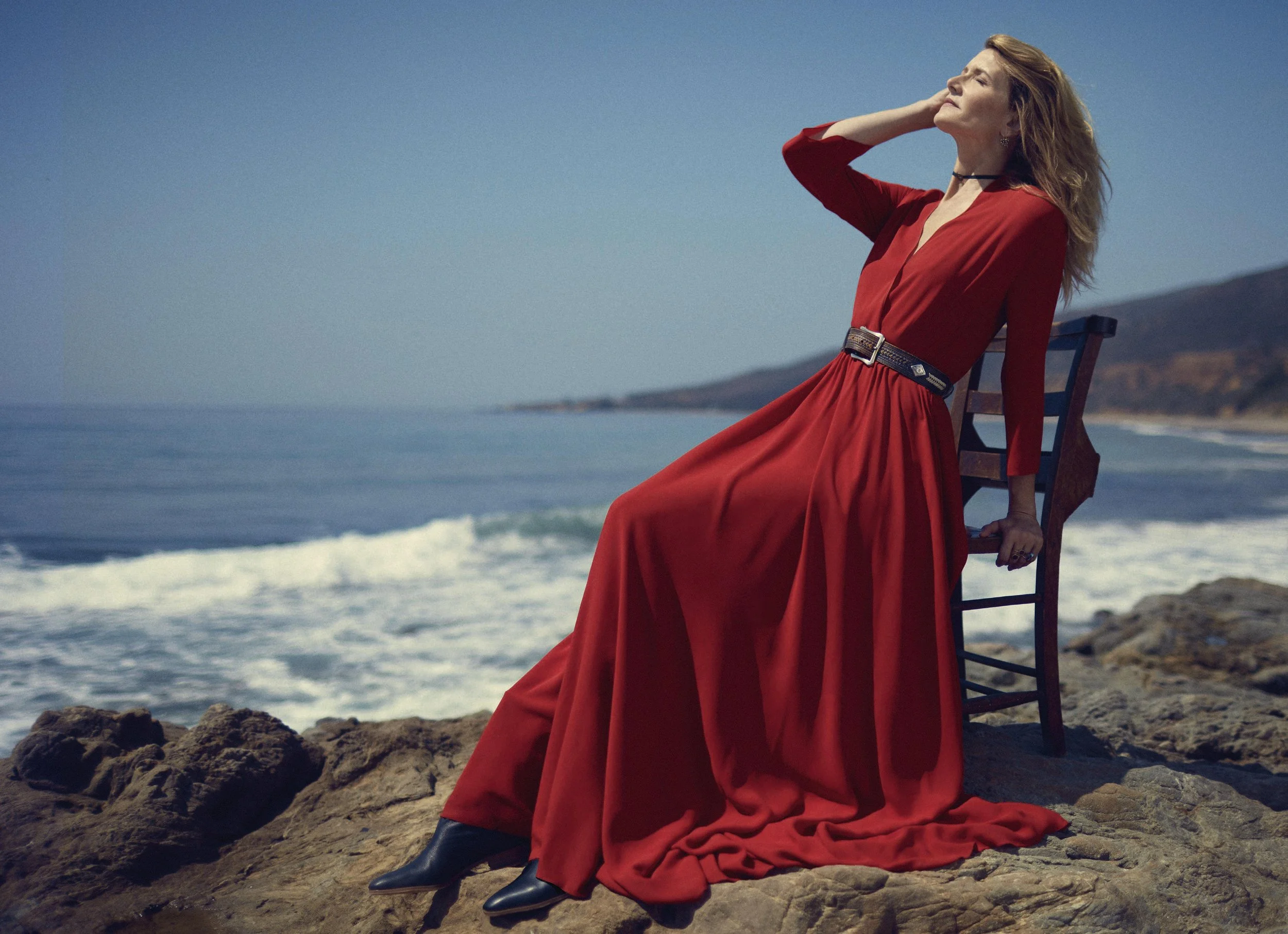Woman in a red dress sitting on a wooden chair on rocky shore, facing the ocean with her hand on her head, ocean waves in the background.