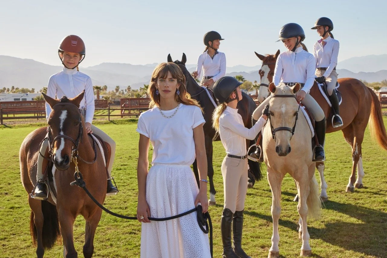 Five women, some on horseback and some on the ground, at a horse ranch with a grassy field and mountains in the background. All are wearing white shirts and helmets.