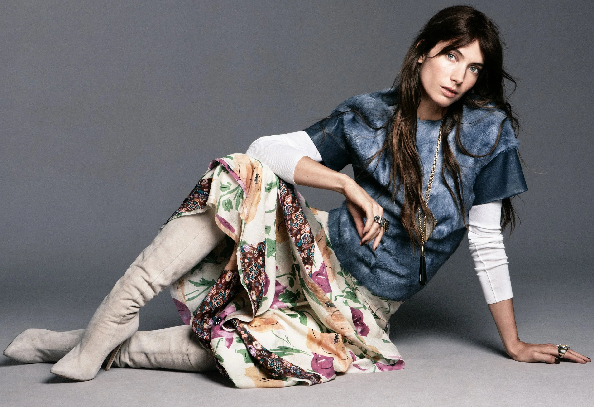 A woman with long brown hair and blue eyes posing on a gray background. She is wearing a layered outfit with a blue and fur top, a white long-sleeve shirt underneath, beige over-the-knee boots, and a colorful floral skirt. She accessorizes with a lon