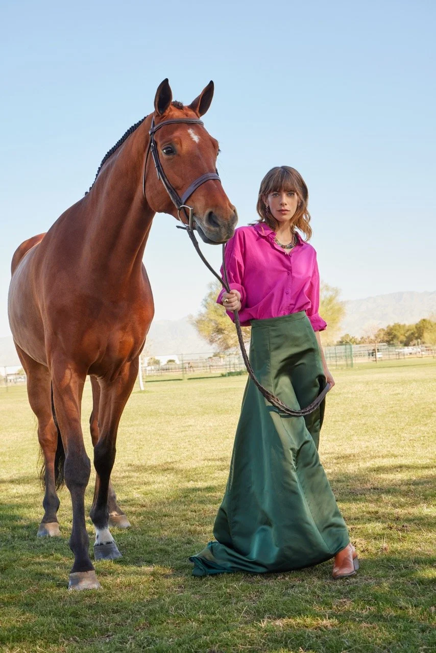 A woman in a pink blouse and green wide-leg skirt holding a brown horse with a bridle on a grassy field under a clear sky.