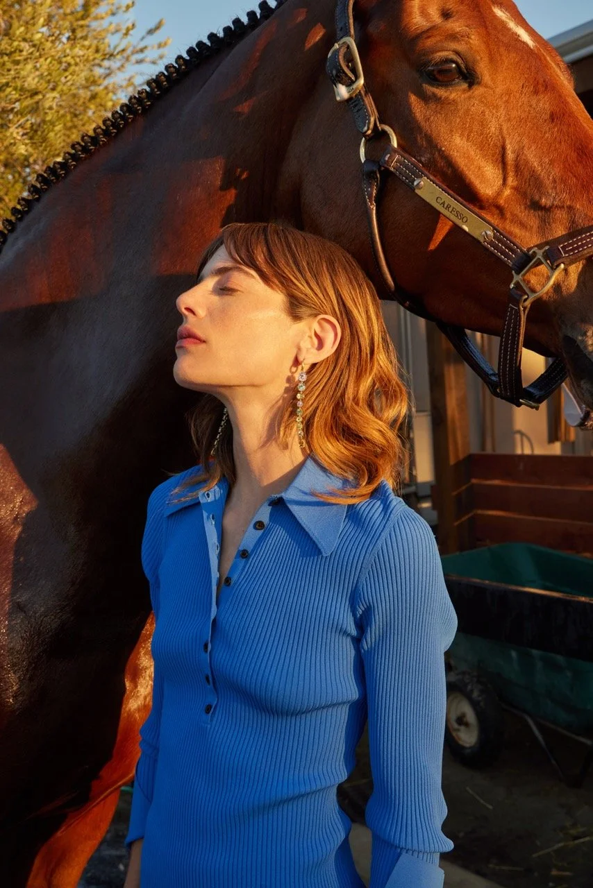 A woman with red hair wearing a blue collared shirt with black buttons stands next to a brown horse with a braided mane, outdoor setting with trees and a building in the background.