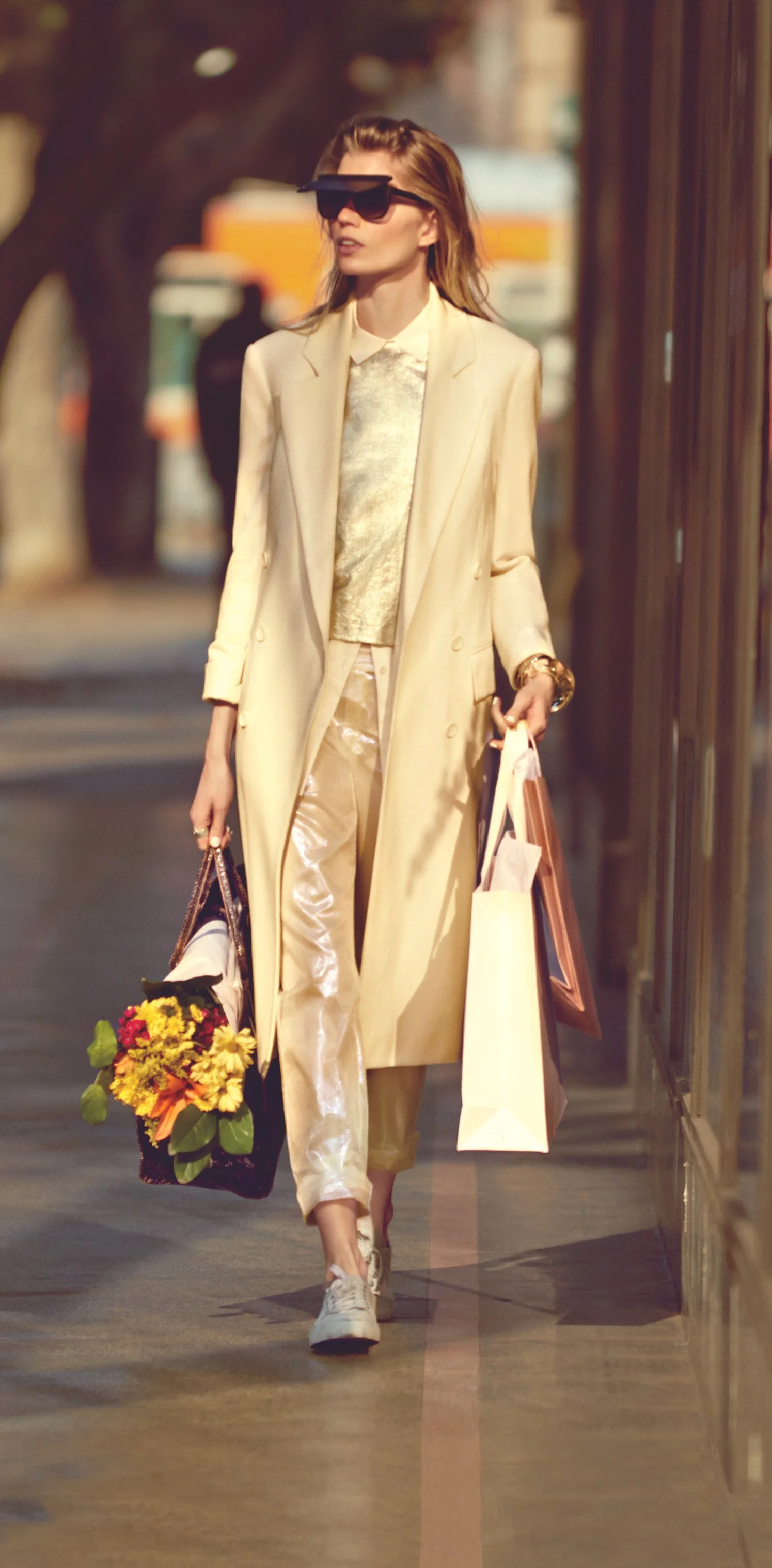 A woman walking on the street carrying shopping bags and a bouquet of flowers, wearing sunglasses, a long beige coat, and stylish clothing.