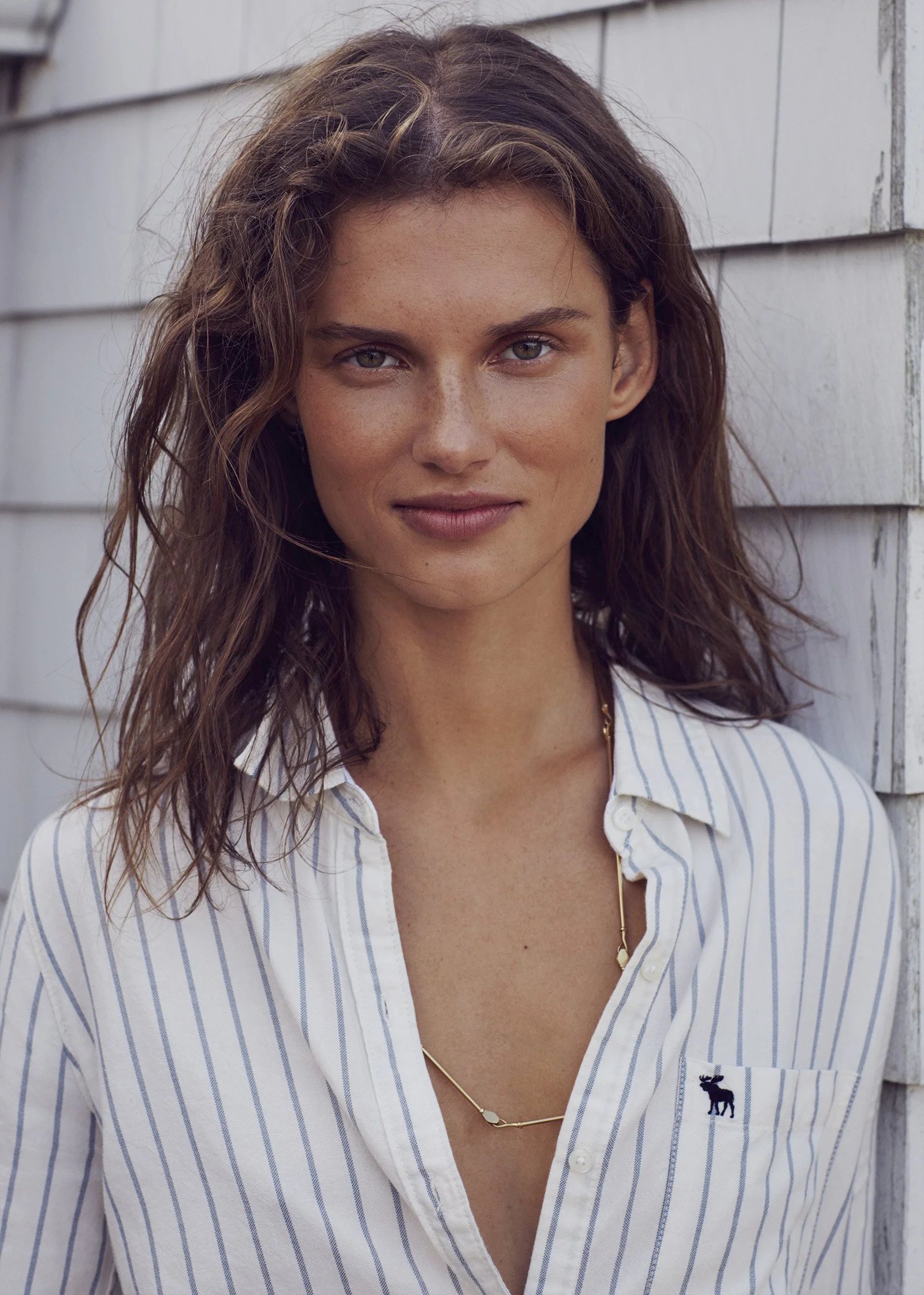 A young woman with wavy brown hair and light skin, wearing a white and blue striped shirt with a small moose logo, standing outdoors against a wooden wall.