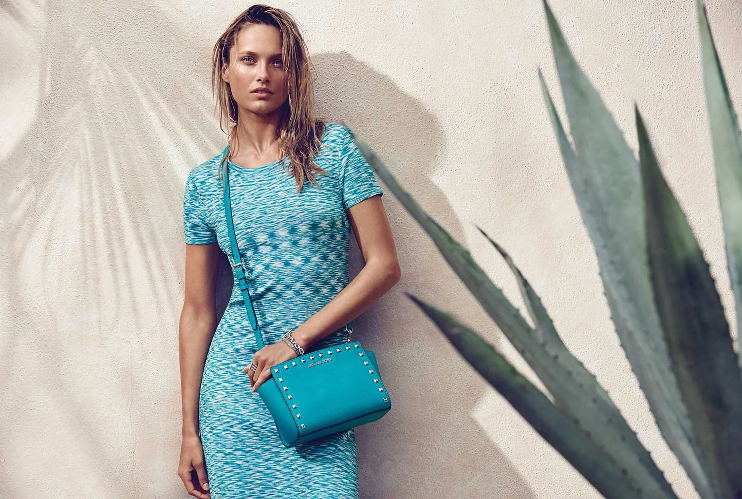 A young woman with wet hair in a blue and white patterned dress stands against a beige wall, holding a blue designer handbag with silver studs, with a large agave plant in the foreground.