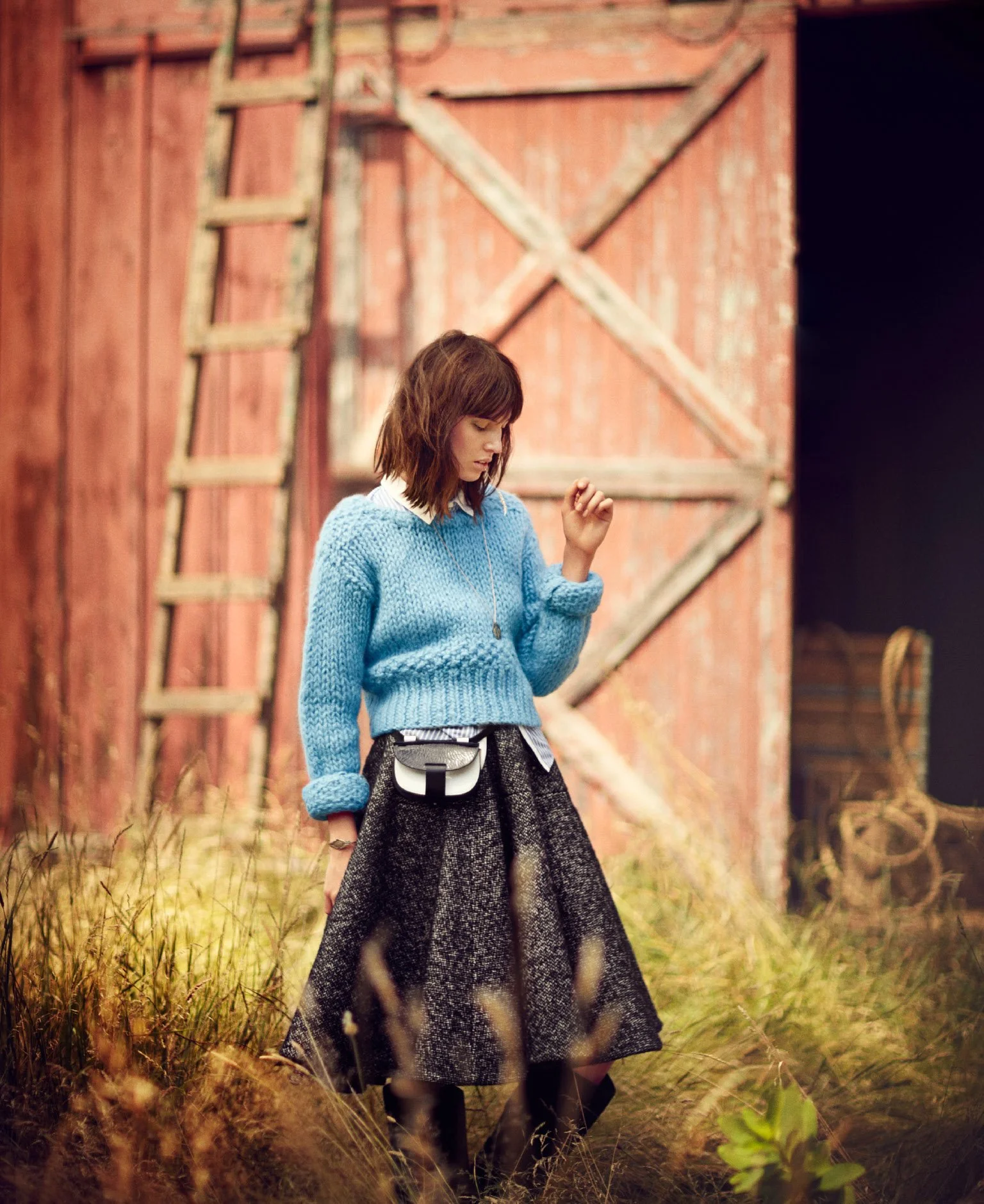 A young woman with shoulder-length brown hair wearing a blue knitted sweater, a black and gray skirt, and black boots, standing outside in front of a rustic red wooden barn with a tall ladder leaning against it, surrounded by tall grass.