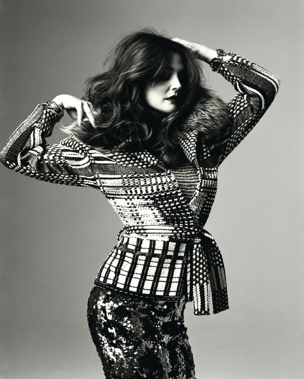 A woman strikes a pose, holding her hair with one hand and her other hand near her head, dressed in a patterned jacket and sequin pants, against a plain background.