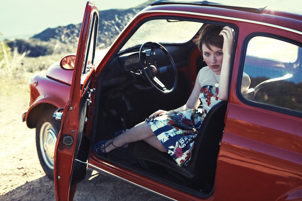 A woman sitting inside a vintage red car with the door open, in a natural outdoor setting with mountains in the background.