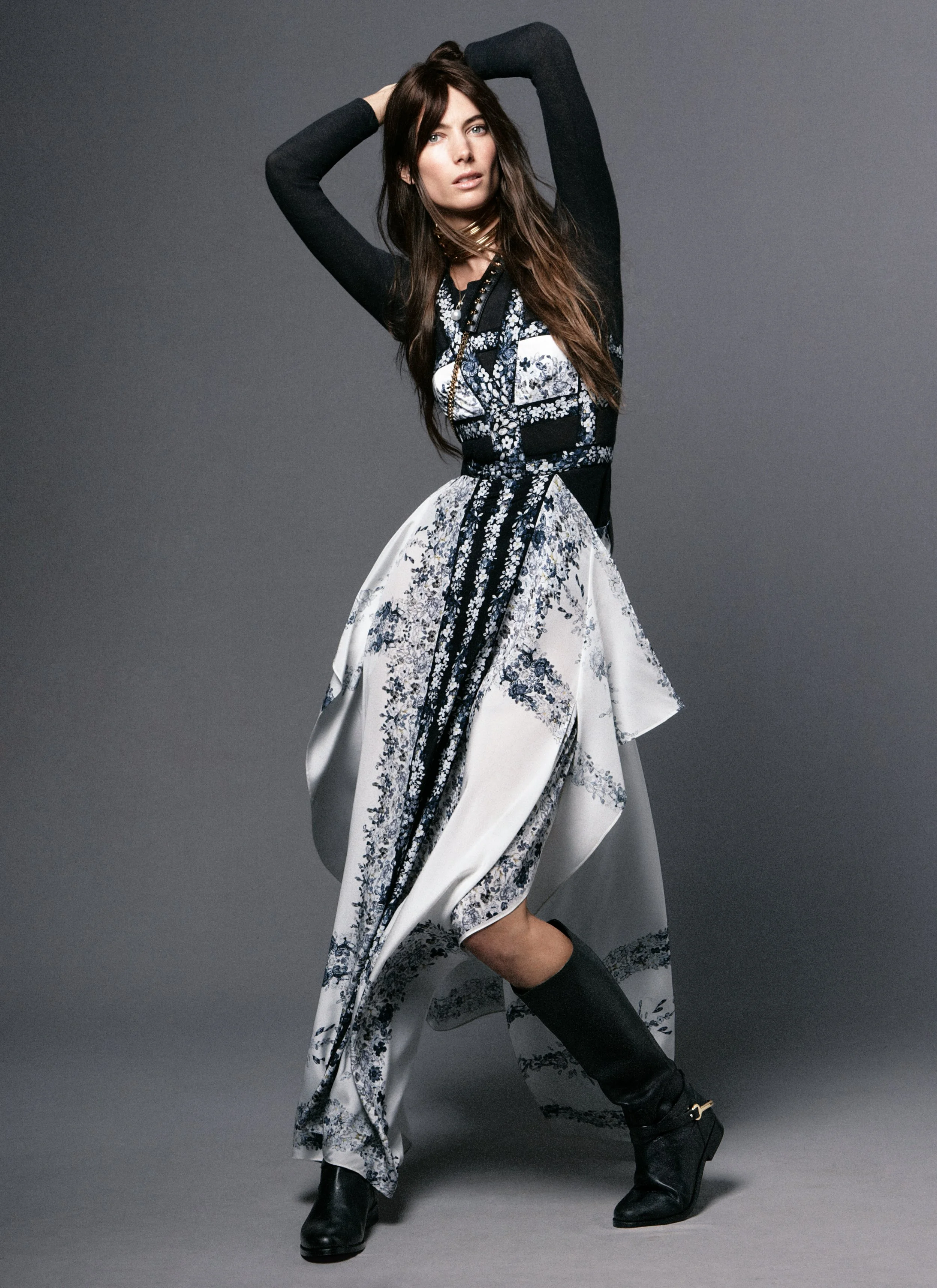 A woman wears a black and white floral-patterned dress with a black long-sleeve top underneath, black boots, and poses with hands behind her head on a gray background.