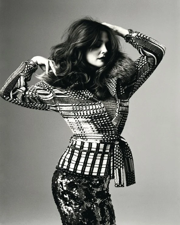 A woman with wavy hair wearing a patterned jacket with a fur collar and shiny pants, posing with one hand on her head in a studio with a plain background.