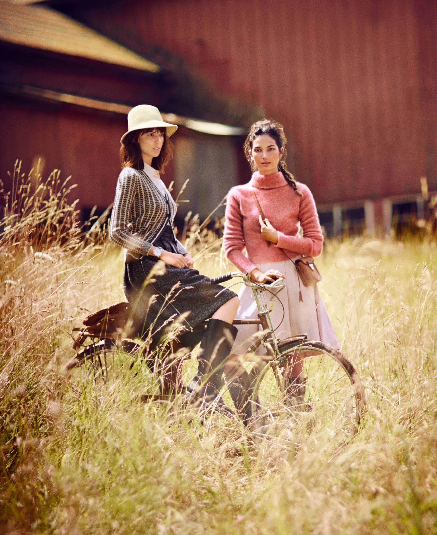 Two women in vintage-style clothing standing outdoors in tall grass, one sitting on a bicycle and the other standing beside it, with a modern building in the background.