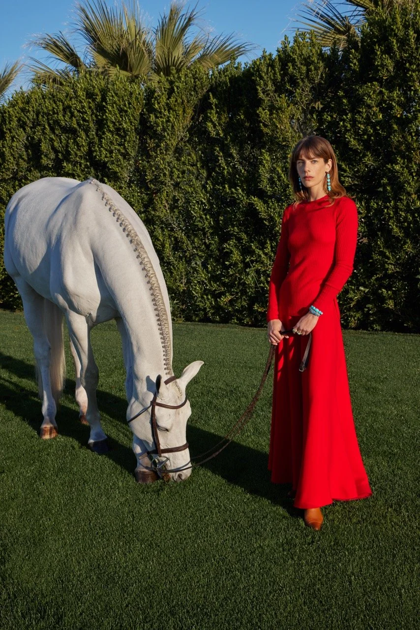 A woman in a long red dress with blue jewelry stands on grass next to a white horse statue in an outdoor setting with green bushes and palm trees.