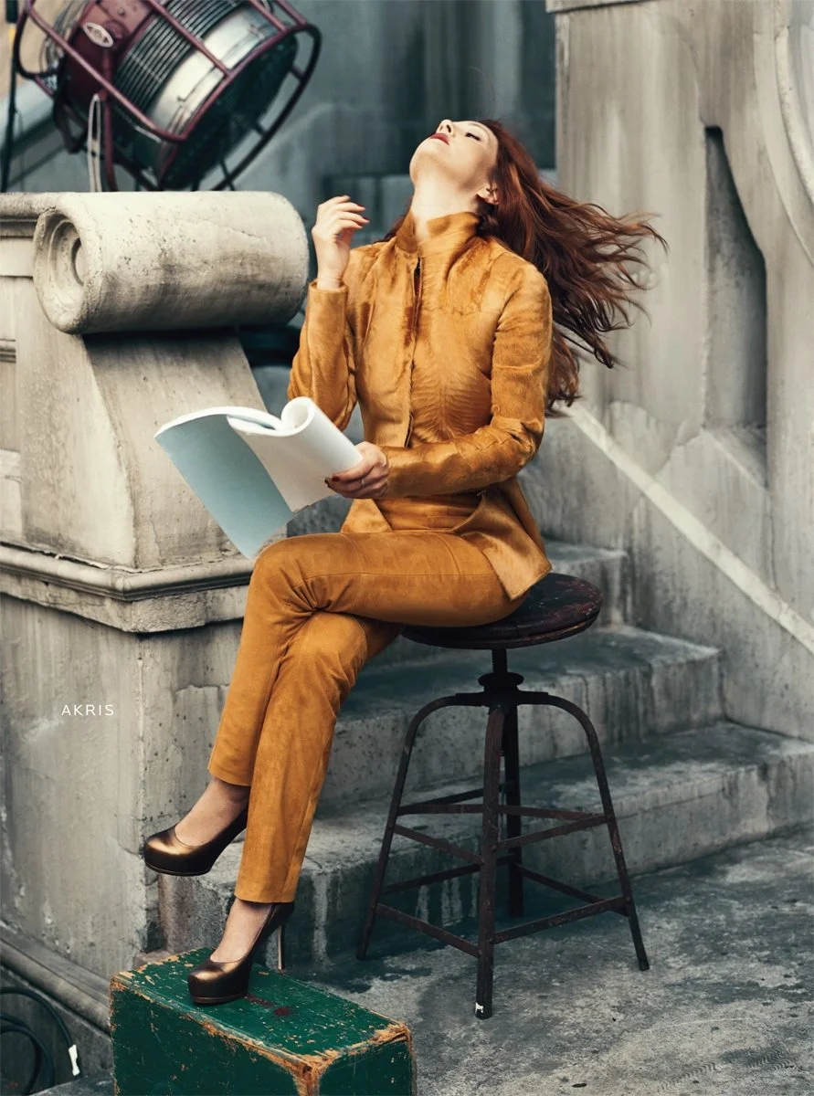A woman with long, flowing red hair is sitting on a stool on a staircase, holding a rolled-up piece of paper in her hand and looking up with her head tilted back. She is wearing a mustard-colored suit and black high heels, with her hair blowing to th