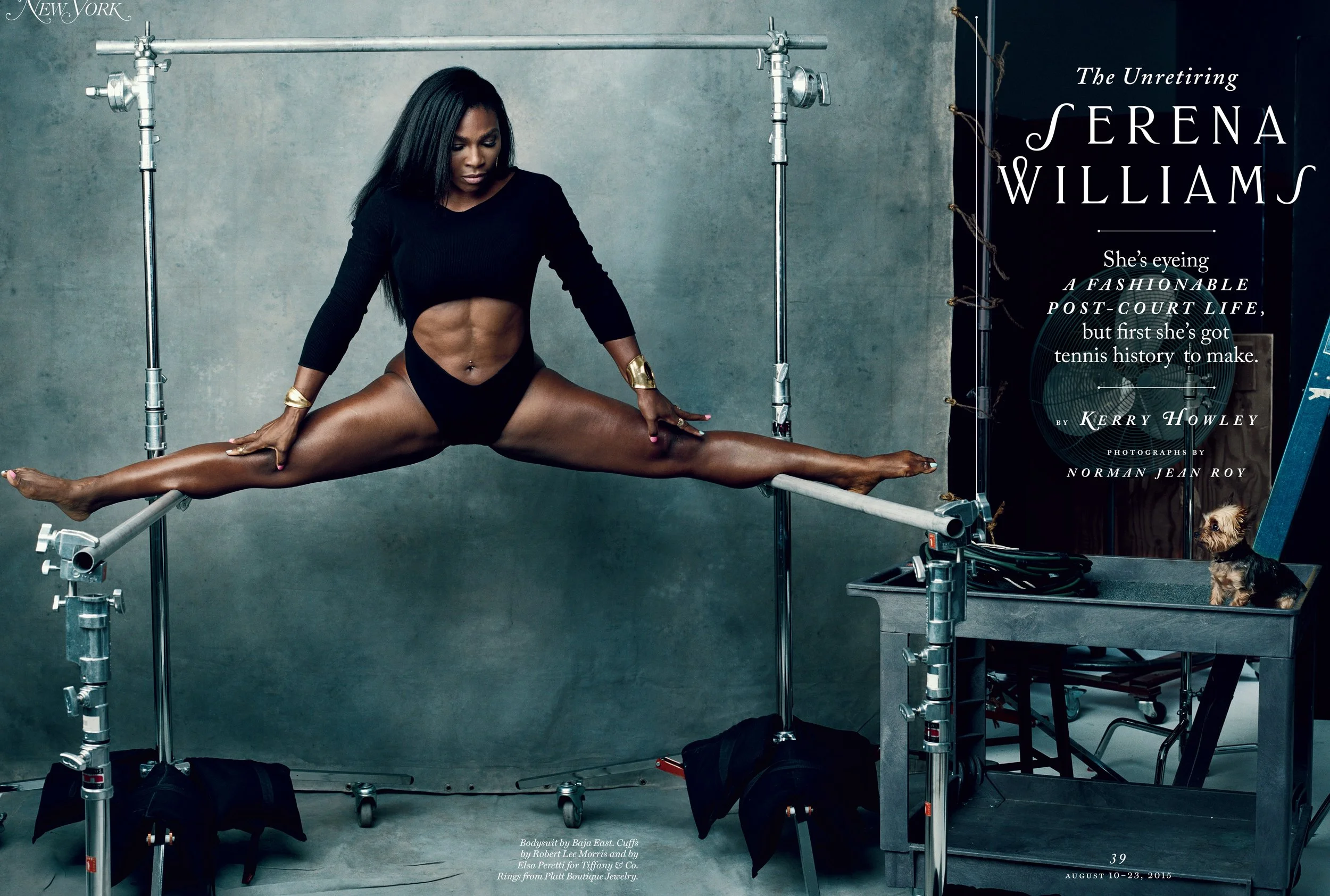 A woman doing a split on a gymnastics beam, wearing a black crop top and black shorts, in a studio with equipment and a small dog sitting on a table beside a ladder.