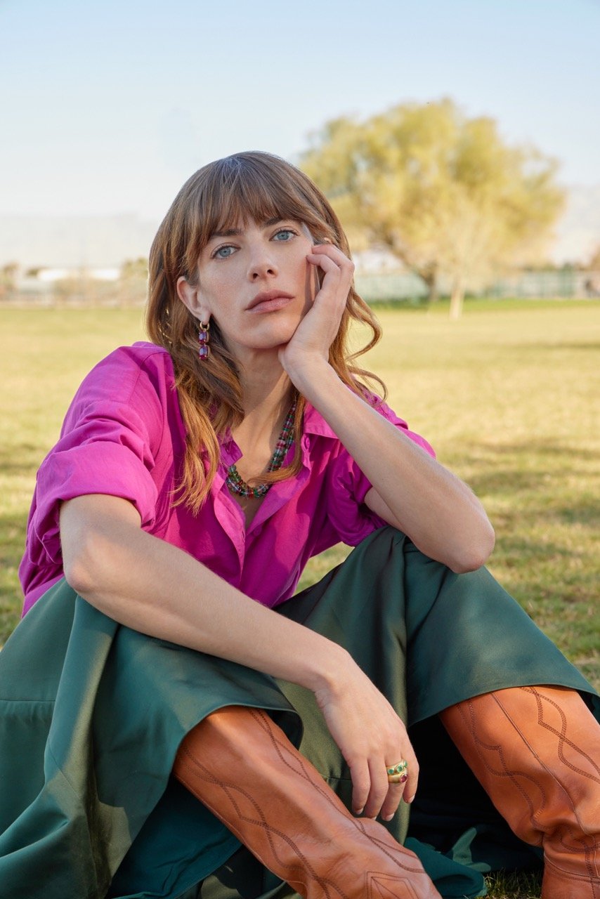 A woman with auburn hair and blue eyes sits on the grass in an open park. She is wearing a pink shirt, green pants, and brown cowboy boots, and she has jewelry including earrings, a necklace, and rings. She rests her head on her hand and gazes though