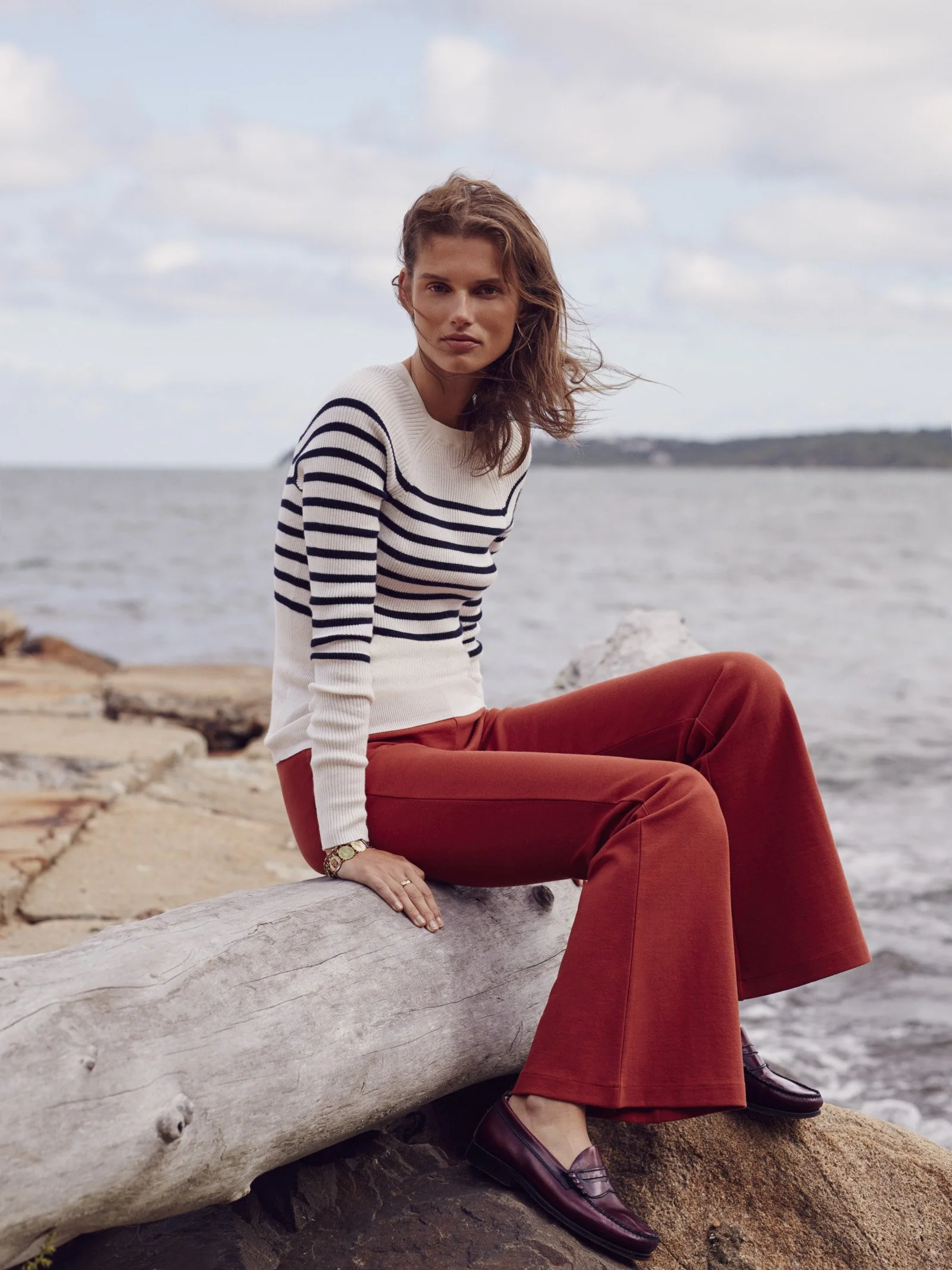 A woman sitting on a large driftwood log near a body of water, wearing a white and navy striped sweater, rust-colored wide-leg pants, and dark loafers. The background features cloudy sky and distant shoreline.