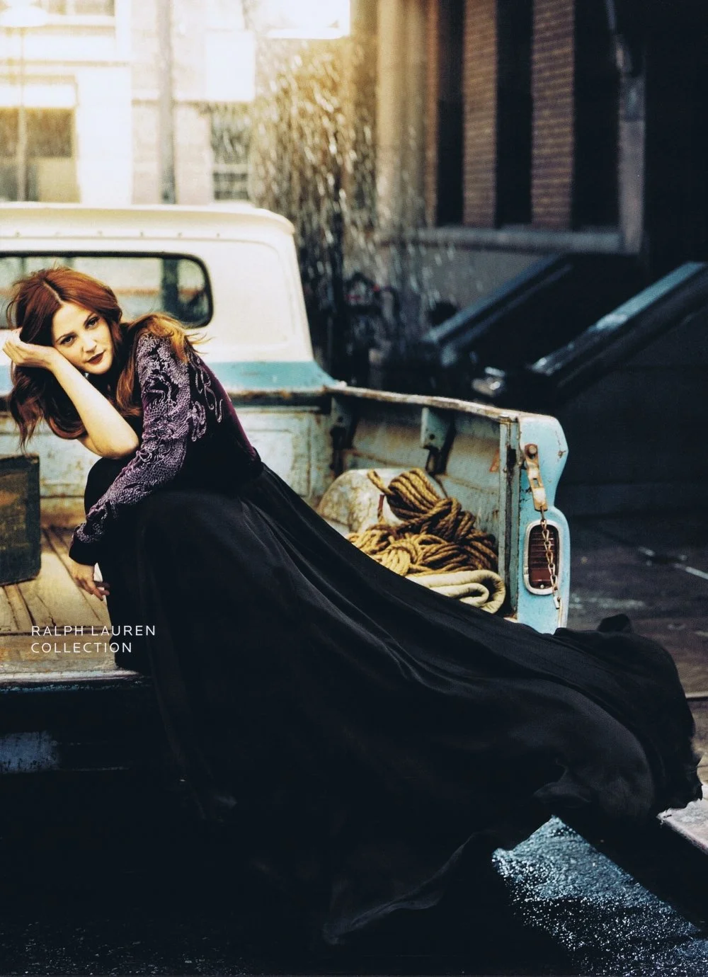 A woman with red hair wearing a black dress with purple lace sleeves, sitting on the side of an old pickup truck with a worn blue bed. The truck contains some ropes. The background shows a brick building and black trash bins. The photo has a warm, sl