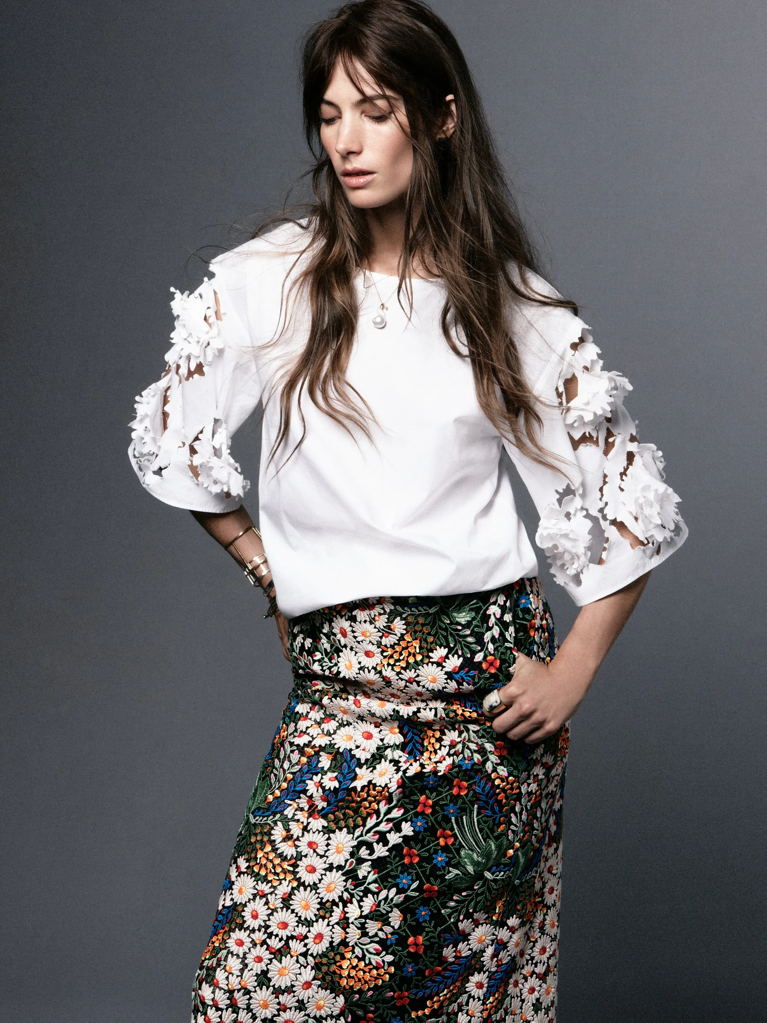 A woman with long brown hair wearing a white blouse with floral cutouts on the sleeves, paired with a colorful floral-patterned skirt, standing against a plain gray background.