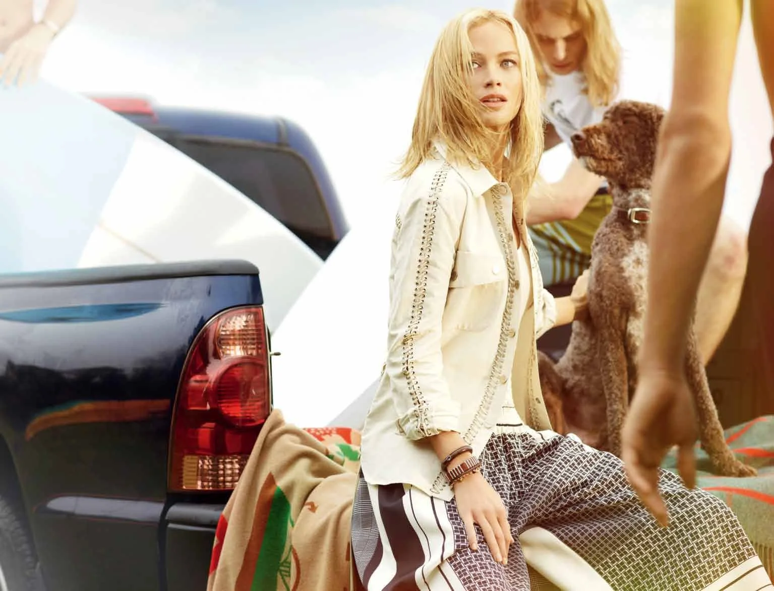Woman sitting on the back of a pickup truck with other people and a dog, outdoors, daytime.