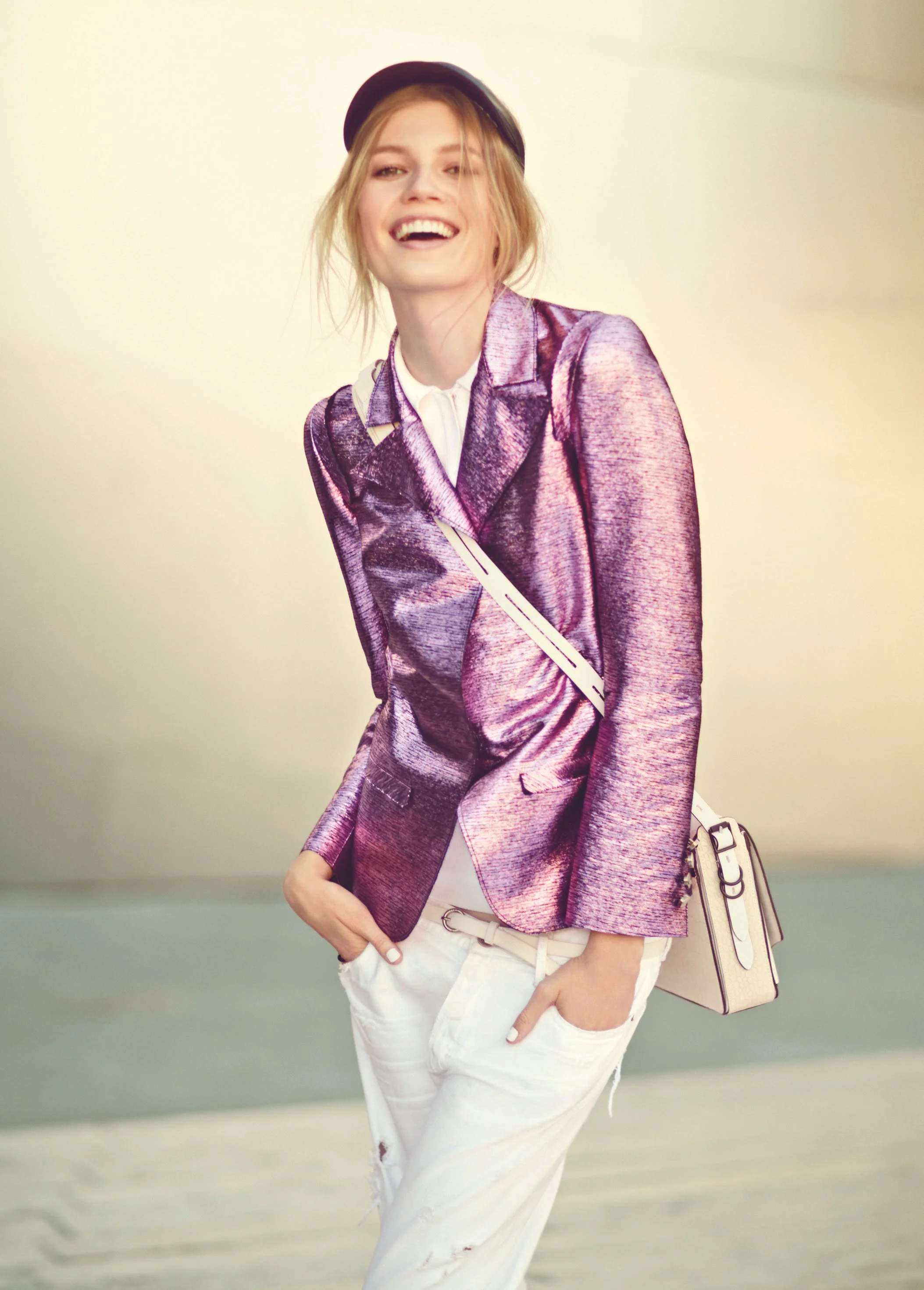 A young woman smiling, wearing a purple shiny blazer, white pants, a white shirt, and a black hat, carrying a beige handbag with white and purple straps.