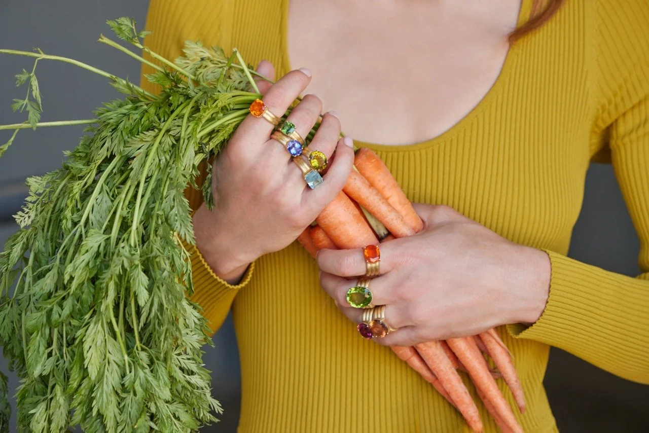 A person wearing a yellow long-sleeve top holding fresh carrots and green leafy vegetables, adorned with multiple colorful gemstone rings.