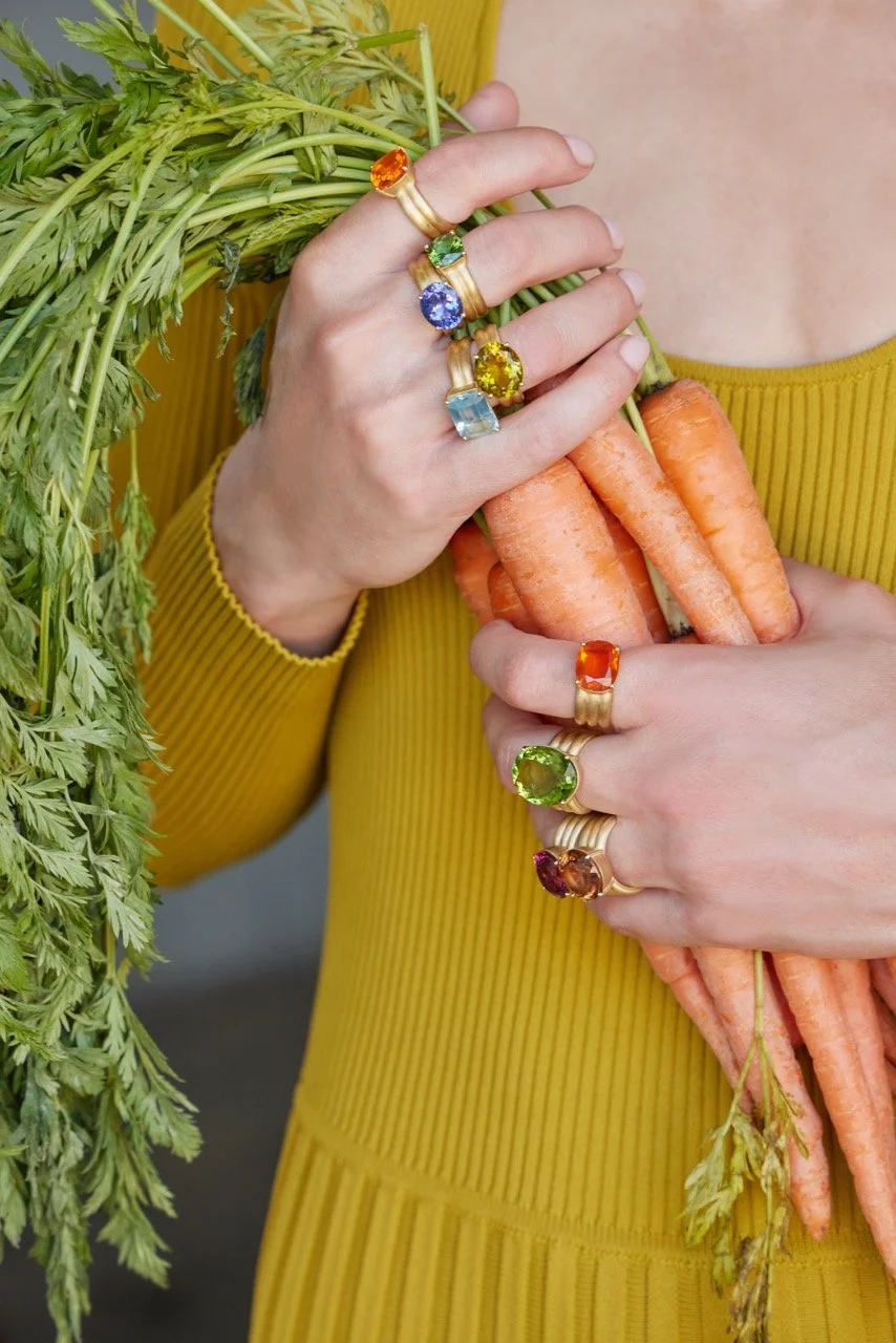 Person wearing a yellow dress holds a bunch of carrots with green tops and showcases multiple colorful gemstone rings.