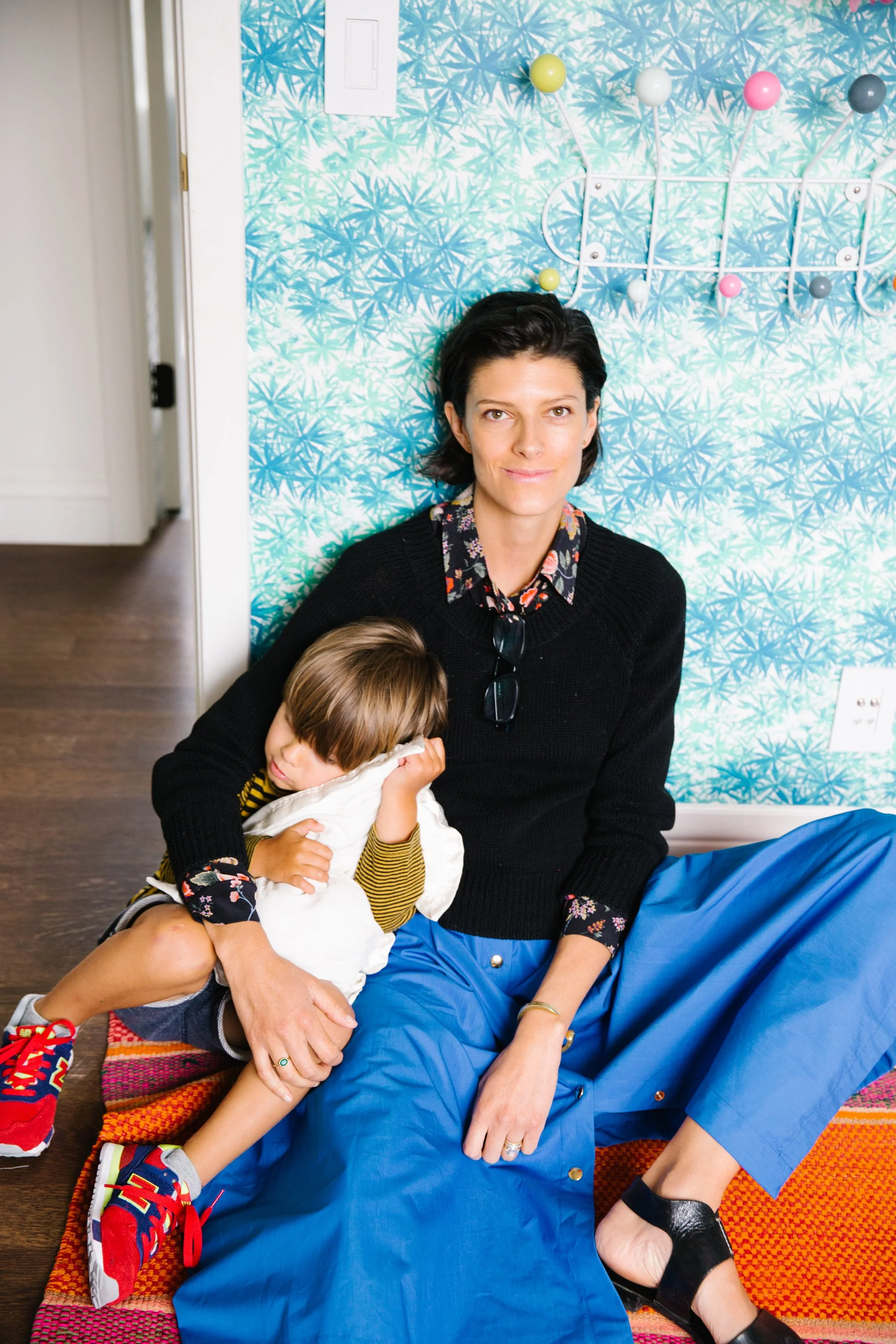 A woman with short dark hair and wearing a black sweater and blue pants sits on a colorful rug, holding a young boy in a white shirt with yellow sleeves who appears to be resting or sleeping against her.