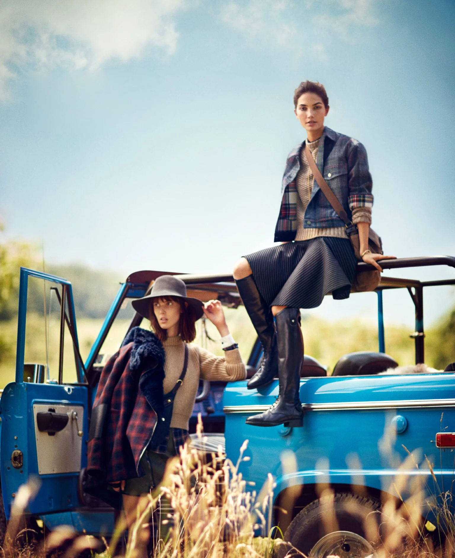 Two women in fashionable fall clothing on and around a vintage blue jeep in an open field during daytime.