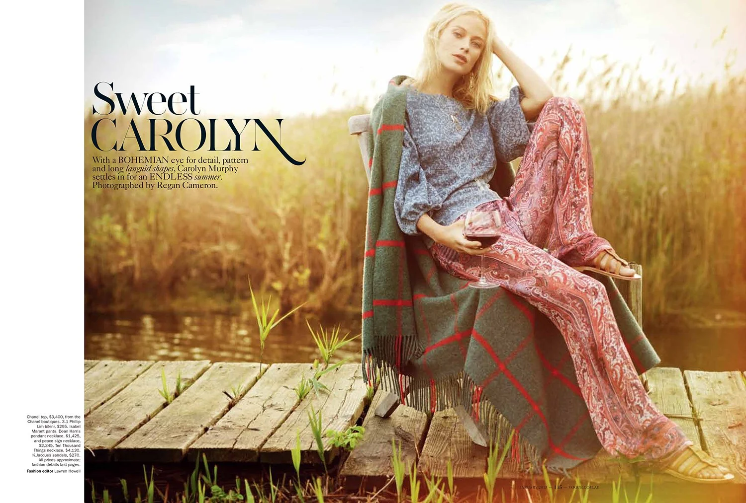 A woman sitting on a wooden dock by a river, holding a glass of red wine, wearing a blue top, red patterned pants, beige sandals, with a green plaid blanket draped over her shoulder, in a natural outdoor setting with tall grasses and water in the bac