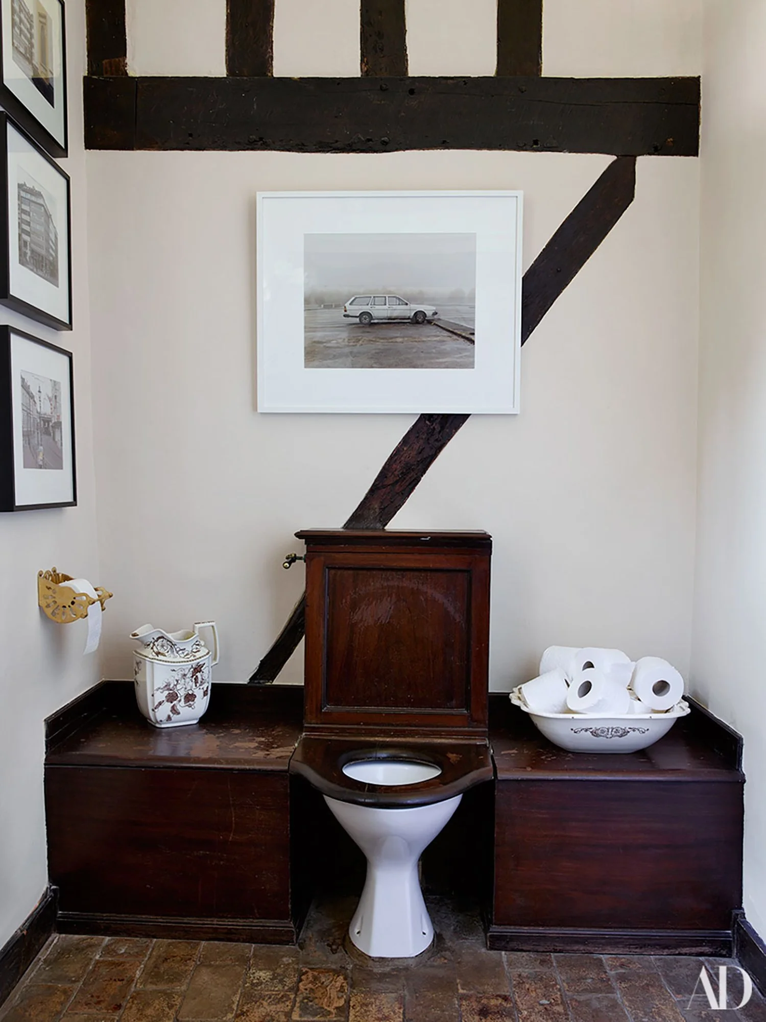 Bathroom with vintage dark wood toilet and fixtures, with a framed photo of a car on the wall, and decorative items including a porcelain pitcher and a bowl of toilet paper rolls.
