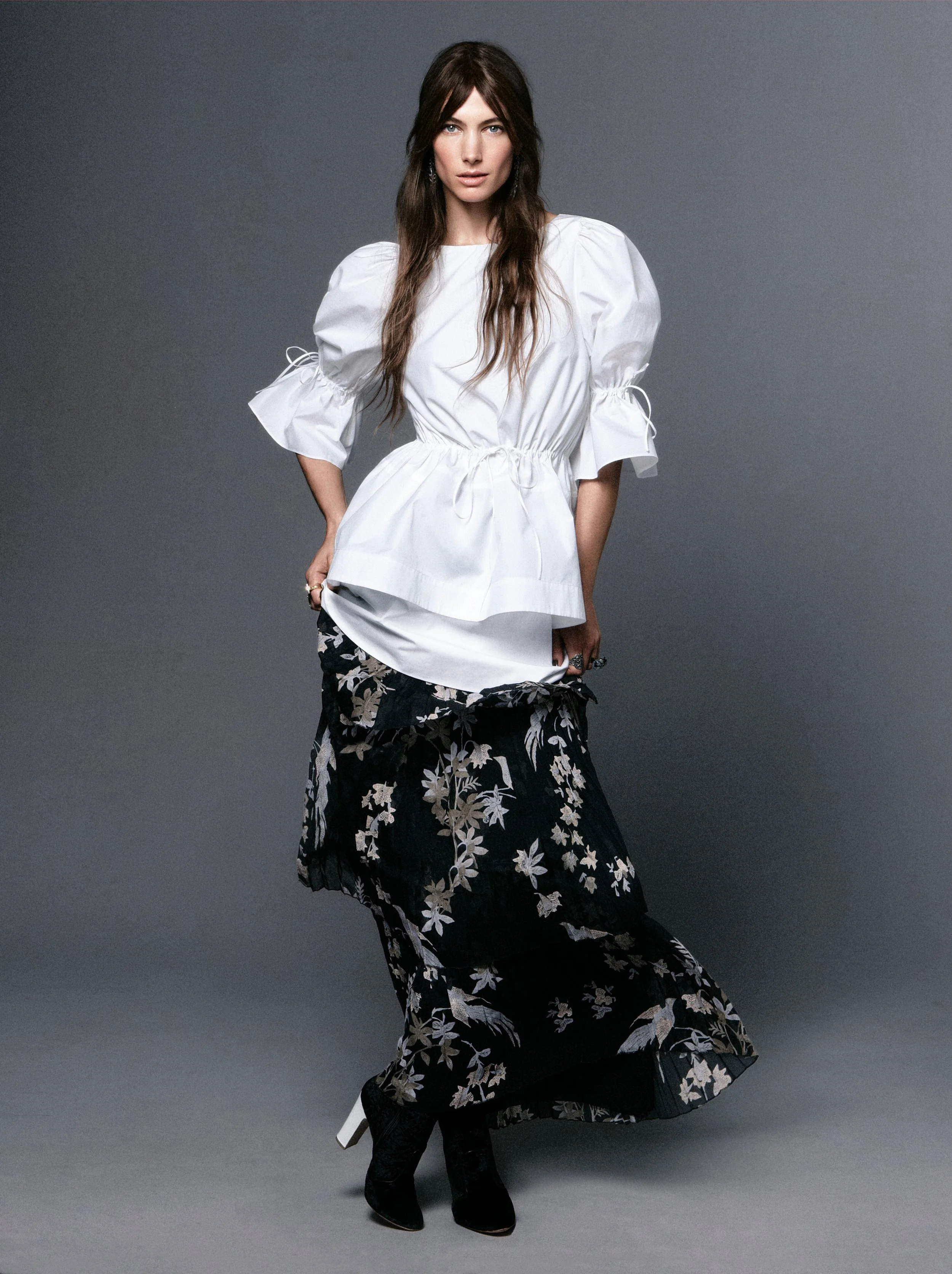 Woman wearing a white top with puffed sleeves and a black, floral-patterned skirt against a gray background.