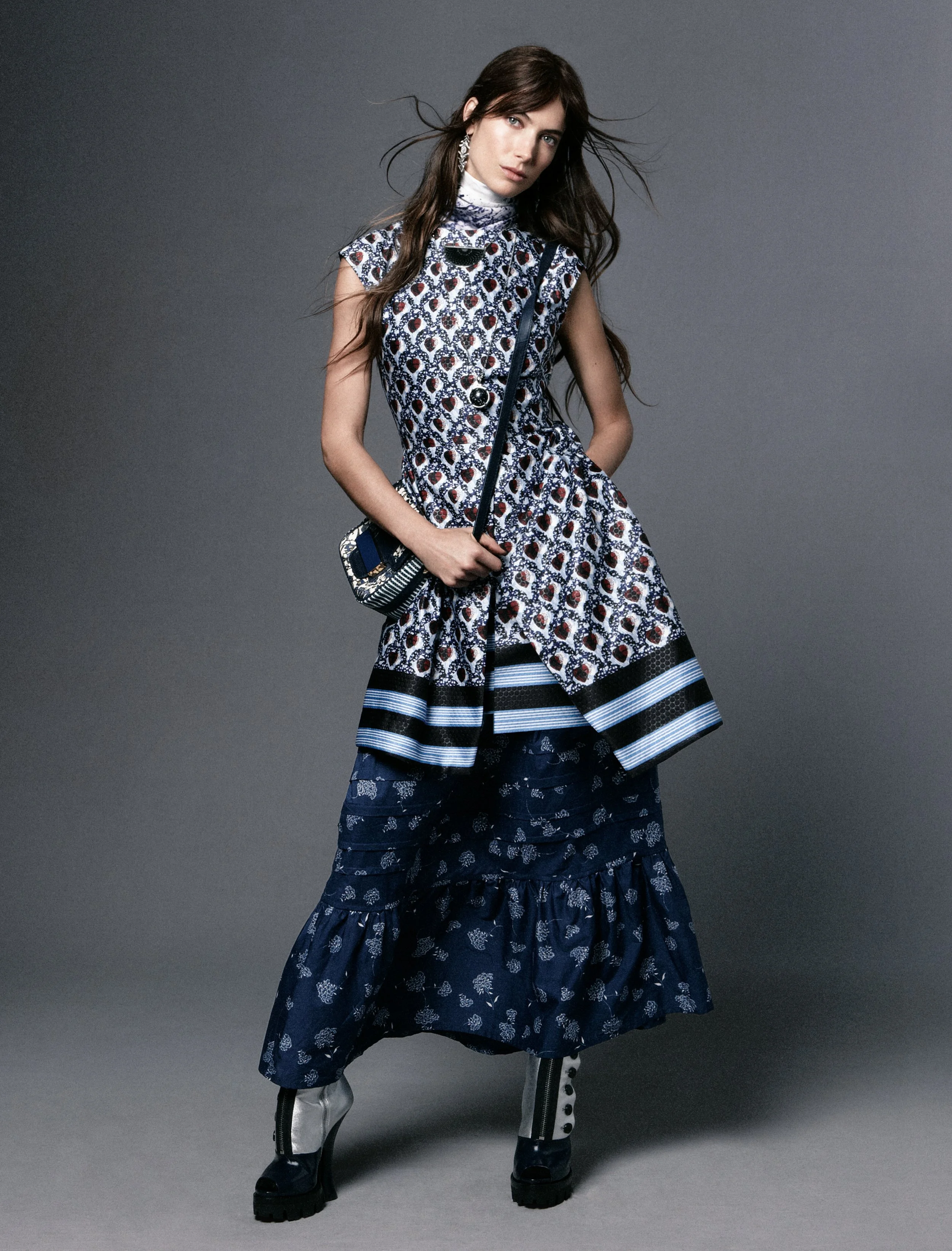 Fashion model wearing a patterned sleeveless top, layered skirt, and boots, holding a small striped handbag against a gray background.