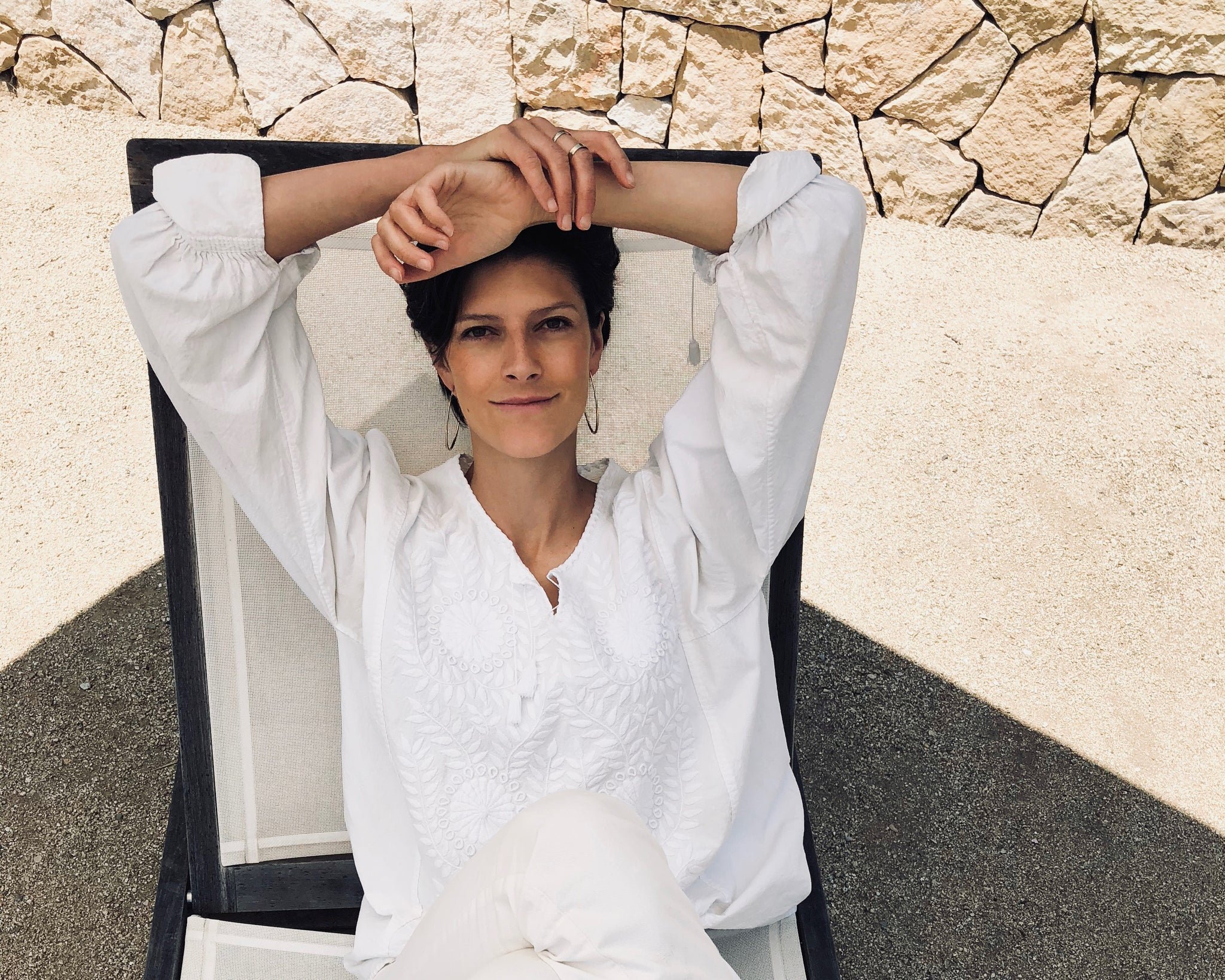 A woman with short dark hair and earrings relaxing on a lounge chair at the beach, wearing a white embroidered blouse, with a sandy and rocky backdrop.