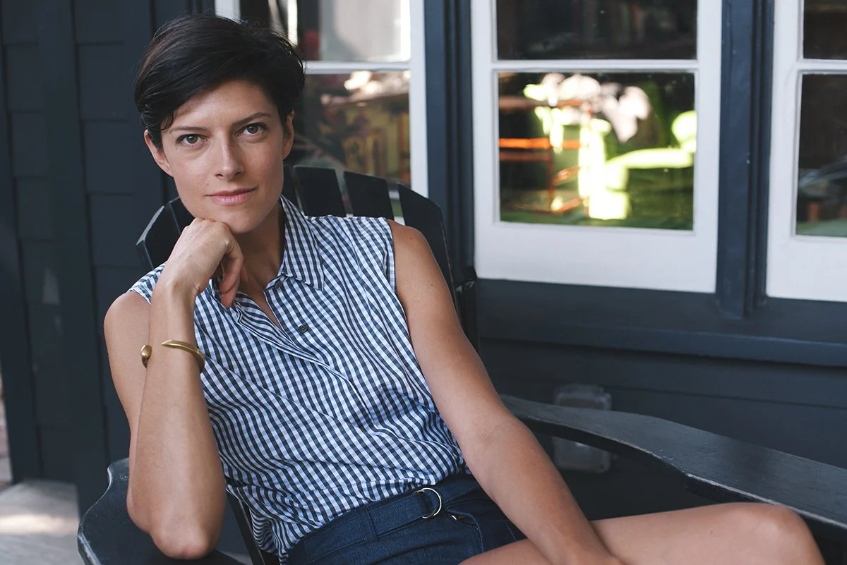 A woman with short dark hair wearing a sleeveless checkered shirt sitting outside on a black chair, resting her chin on her hand, with a house with dark exterior and windows in the background.
