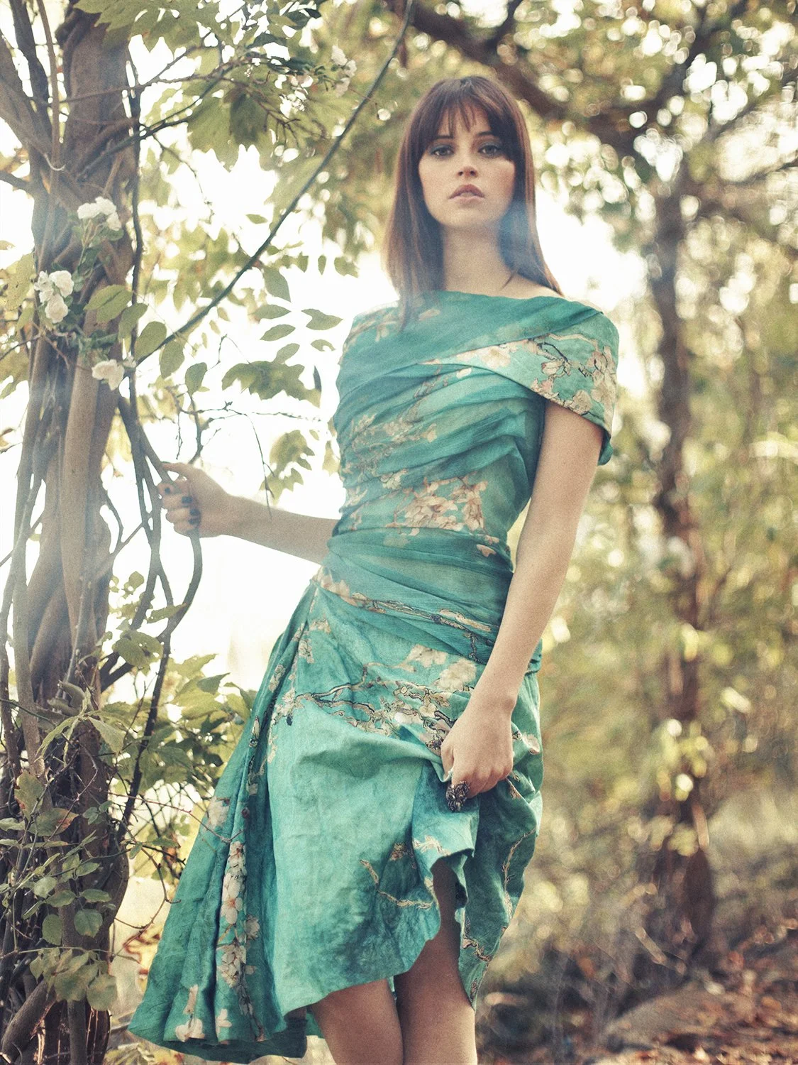 A woman in a teal patterned dress stands outdoors near a tree, holding a branch with leaves, with sunlight filtering through the background foliage.