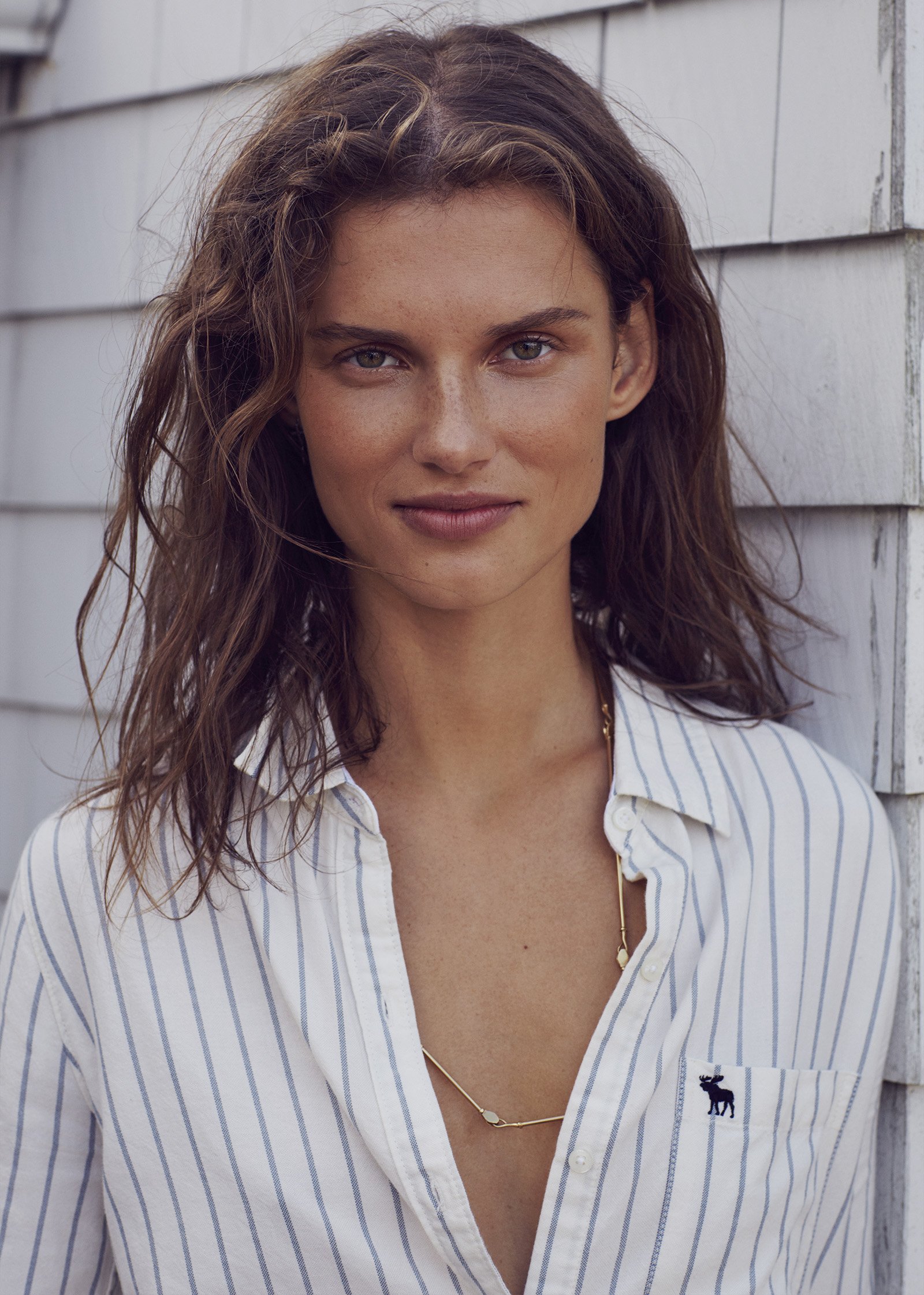 A woman with wavy brown hair and light skin standing outdoors against a white wooden wall, wearing a white and blue striped button-up shirt with a small dark embroidered moose on the chest and gold necklaces.