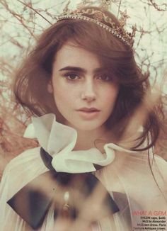 Young woman with dark hair and a tiara, wearing a white bow in her hair and a light-colored dress, standing outdoors with blurred autumn leaves in the background.