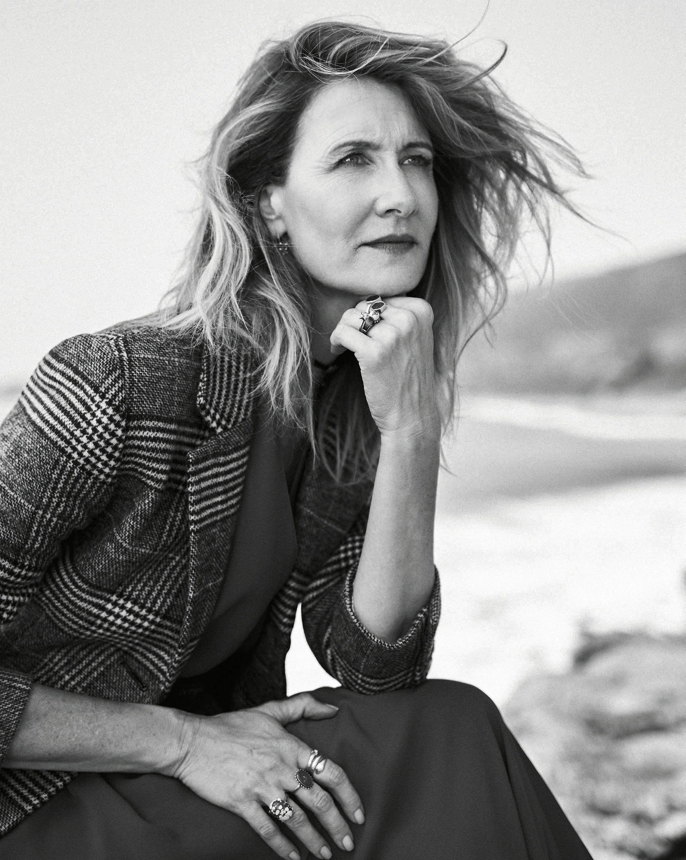 A woman with wavy hair, wearing a checkered blazer and multiple rings, sitting outdoors with a thoughtful expression, resting her chin on her fist and looking into the distance.