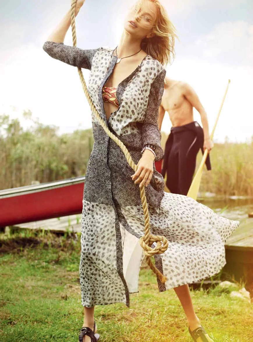 A woman with blonde hair wearing a patterned dress holding a thick rope outdoors near a boat, with a shirtless man holding a paddle in the background.