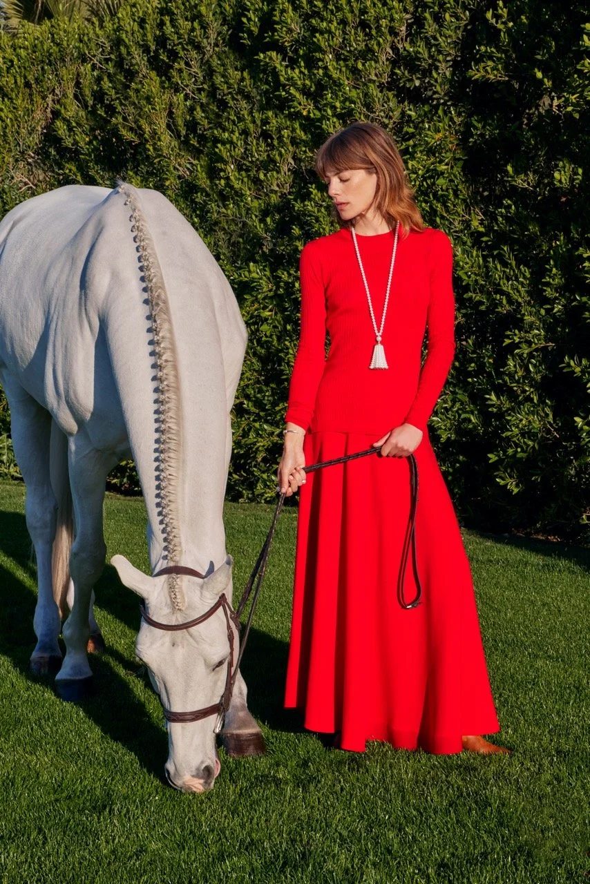 A woman wearing a long red dress and a long necklace with a pendent is holding a leash attached to a white horse that is grazing on the grass. The woman has shoulder length brown hair and is standing outside in front of a green hedge.
