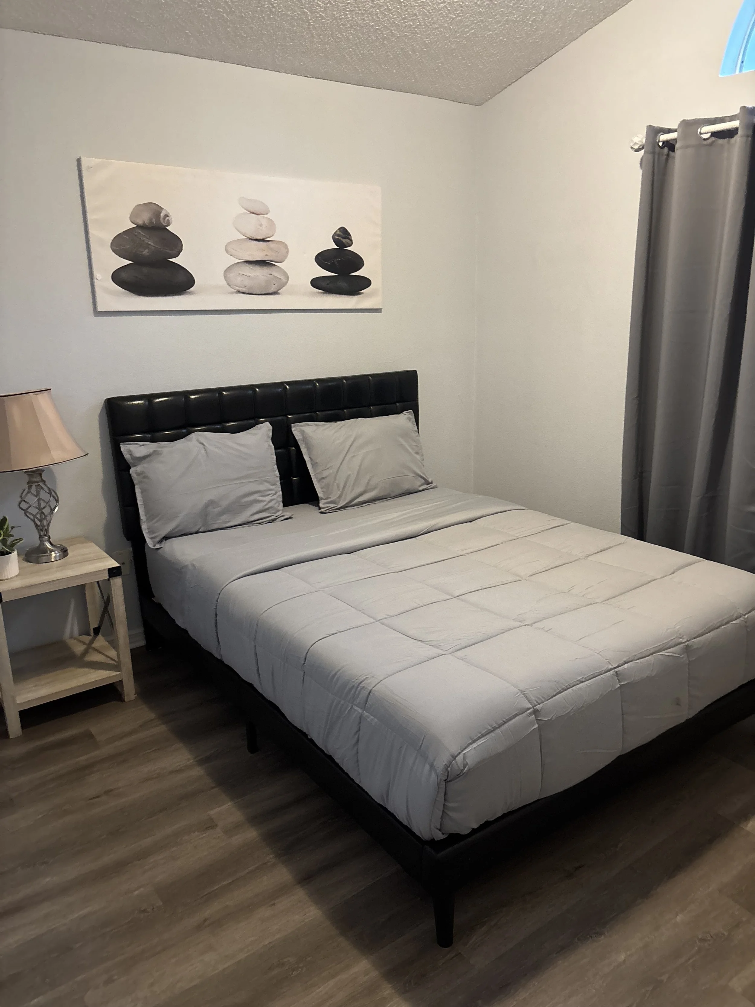 A bedroom with a bed that has gray pillows and a beige comforter, a nightstand with a lamp and small plant, gray curtains, and a wall hanging of stacked stones.