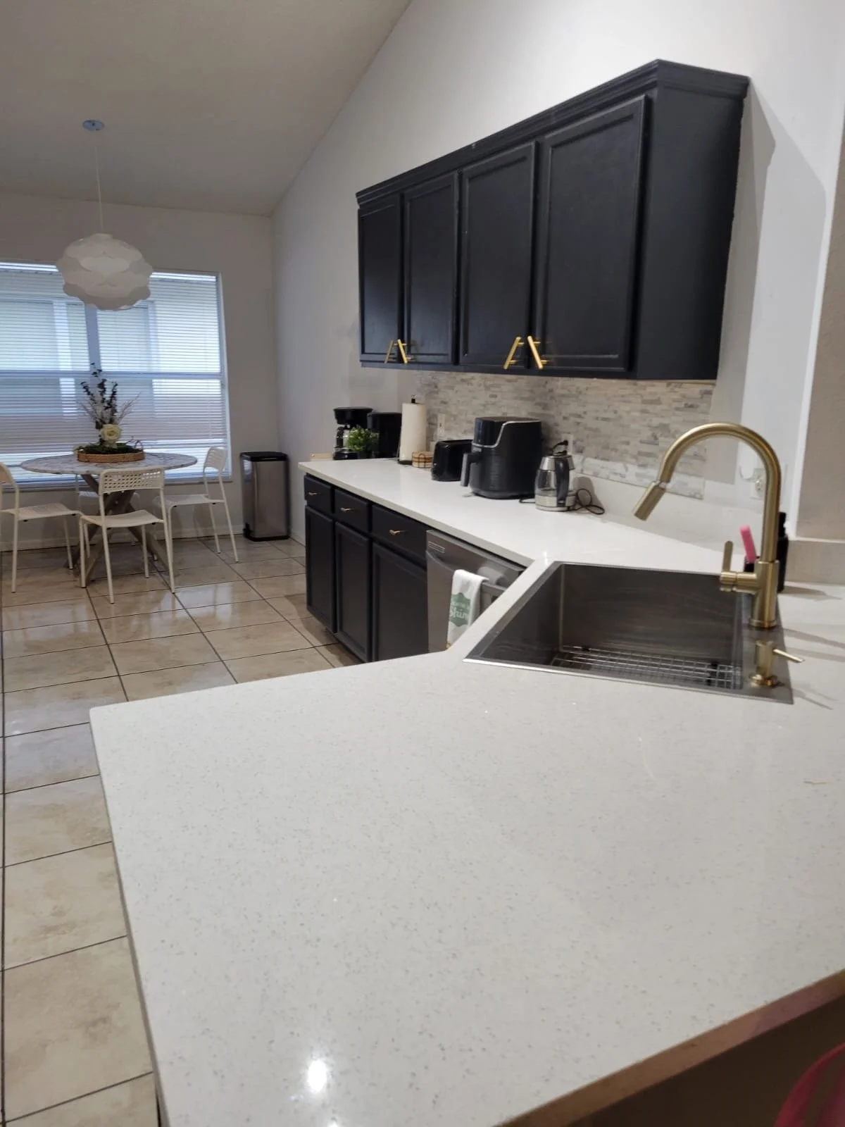 Kitchen with white speckled countertop, black cabinets, a gold faucet in a stainless steel sink, small appliances on the counter, and a dining area with a round table and chairs near a window with blinds.