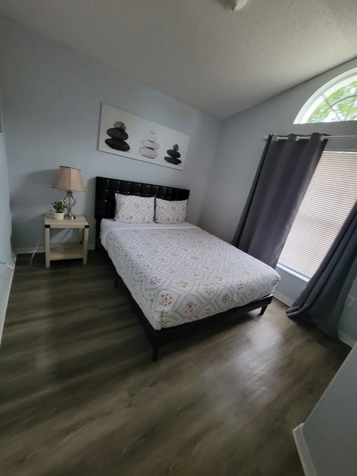 A bedroom with a bed that has a black upholstered headboard, white bedding, and two decorative pillows. To the left is a small wooden nightstand with a lamp and a potted plant. There is a window with partially closed shades and dark gray curtains. A 