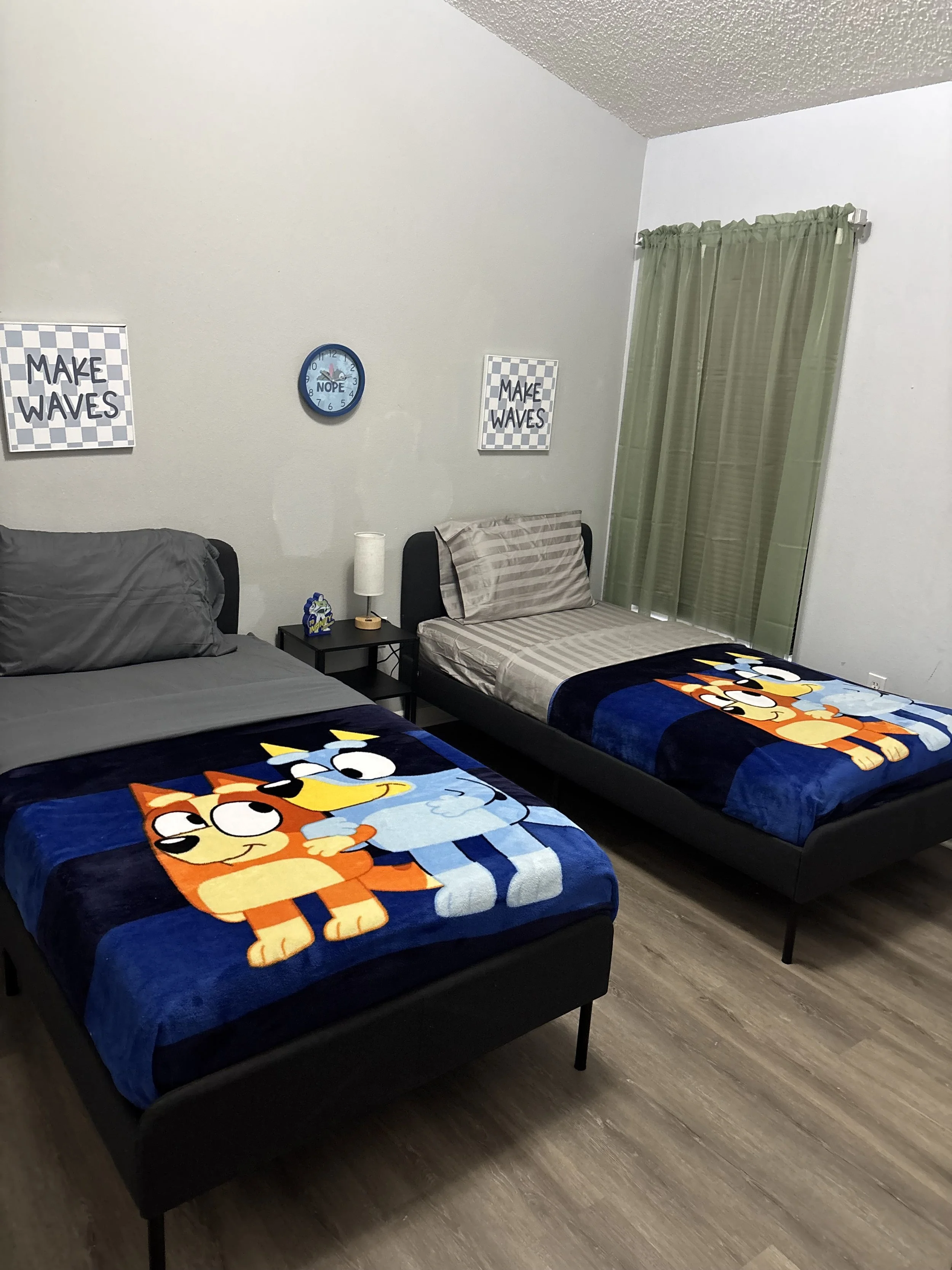 A bedroom with two beds, Garfield and Odie themed blankets, green curtains, and wall decor including a clock and posters reading "Make Waves."
