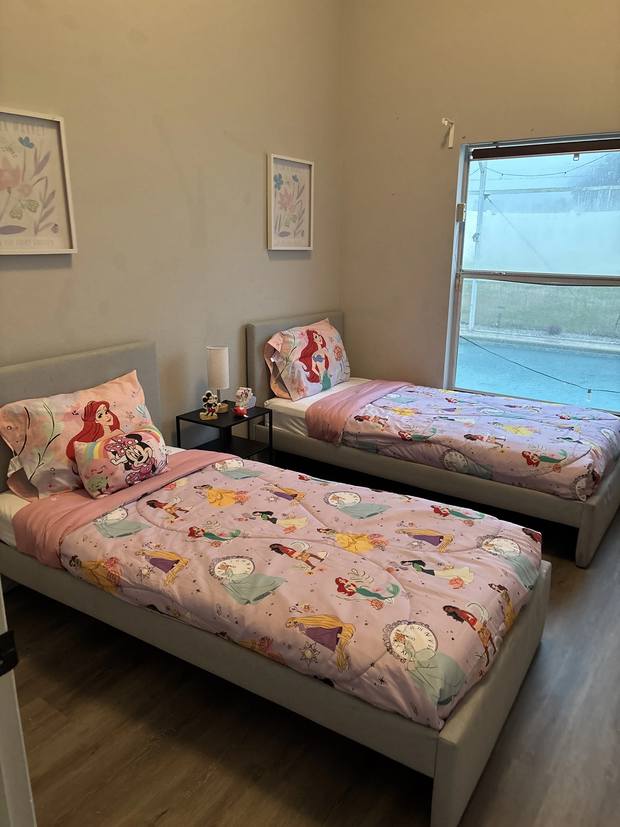 Bedroom with two twin beds featuring Disney princess-themed bedding and pillows, a black nightstand with a lamp and figurines, and a window with a view of the outside.