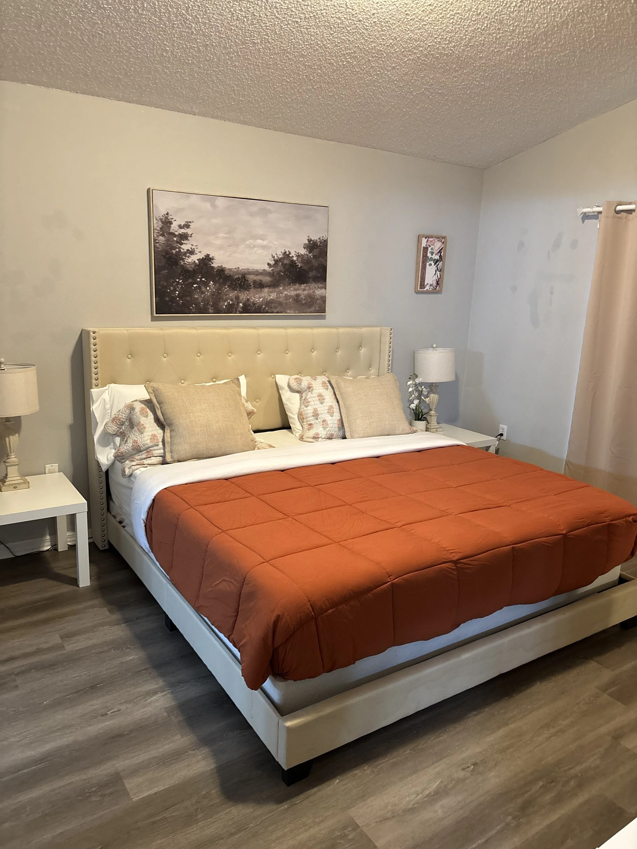 A neatly made bed with a beige tufted headboard, orange comforter, multiple pillows, flanked by white nightstands with lamps, in a bedroom with light-colored walls, framed artwork, and wood-like flooring.