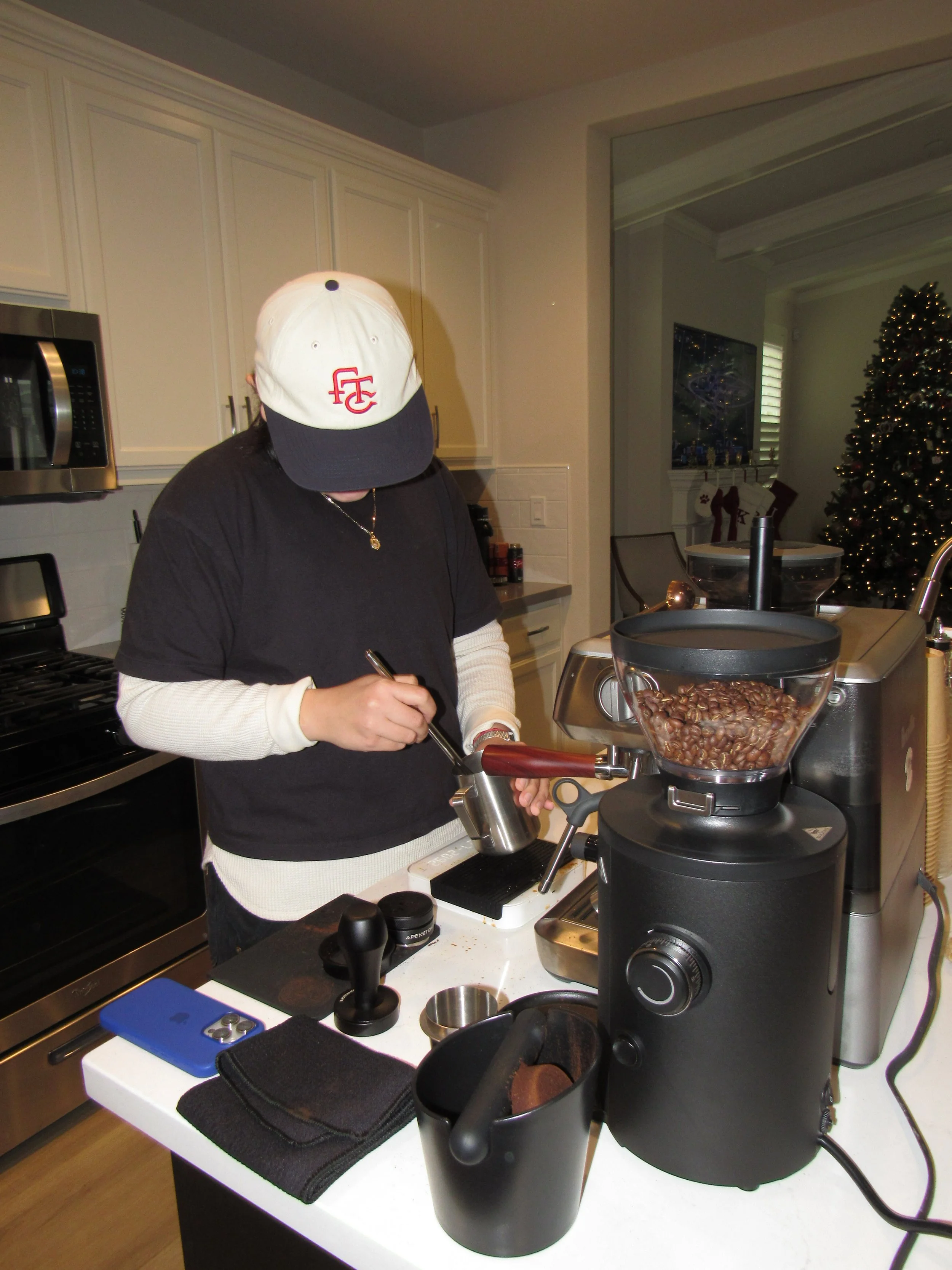 A person wearing a white cap with red initials, black shirt, and white long sleeves, standing at a kitchen counter and preparing coffee with a coffee grinder filled with coffee beans.