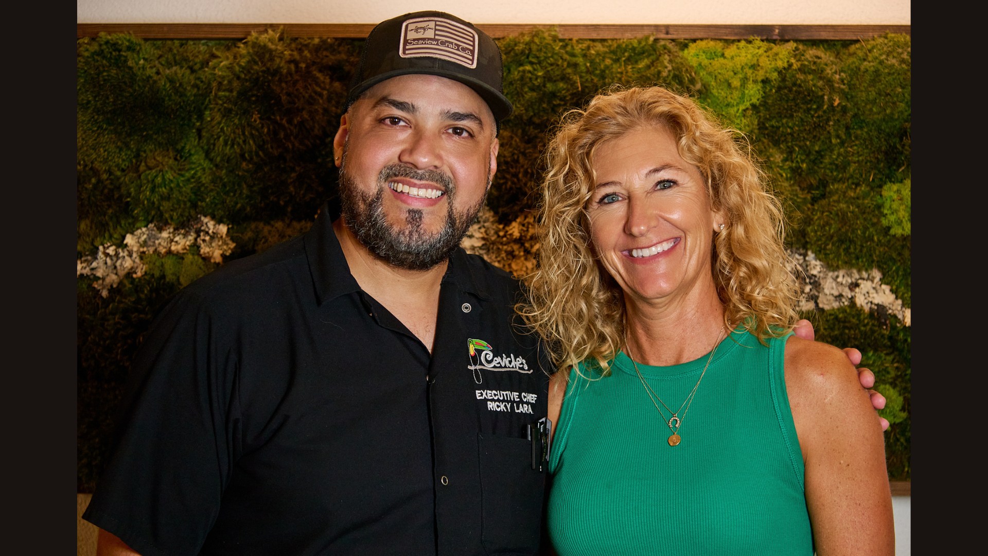 A man with a dark beard and mustache wearing a black cap and black chef's shirt standing next to a woman with curly blonde hair wearing a green sleeveless top. They are both smiling and posing for the camera, with a textured green background.
